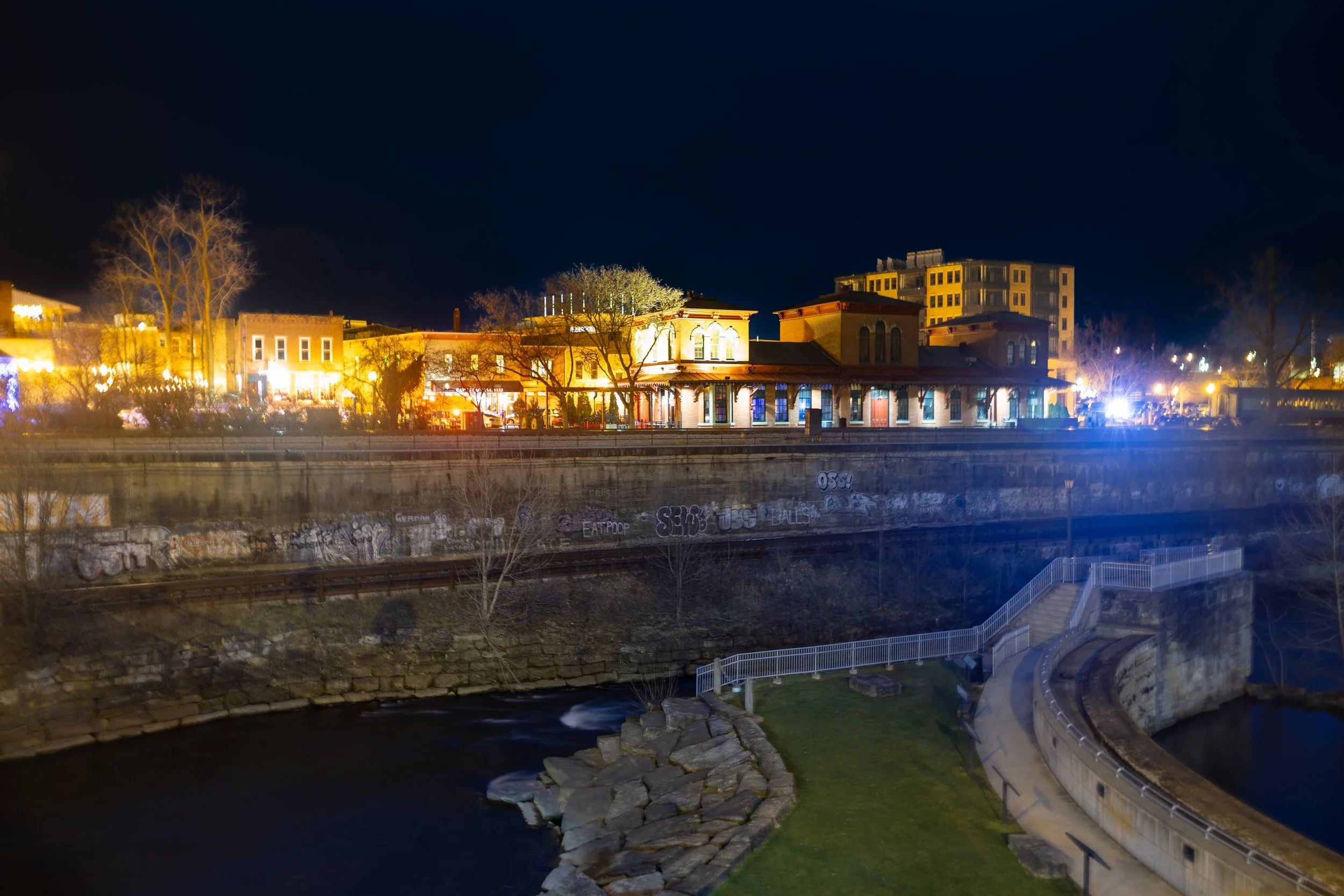 Night view of a cityscape with illuminated buildings across a river, including graffiti on the wall along the water, with a curved walkway and stairs leading down to the riverbank.