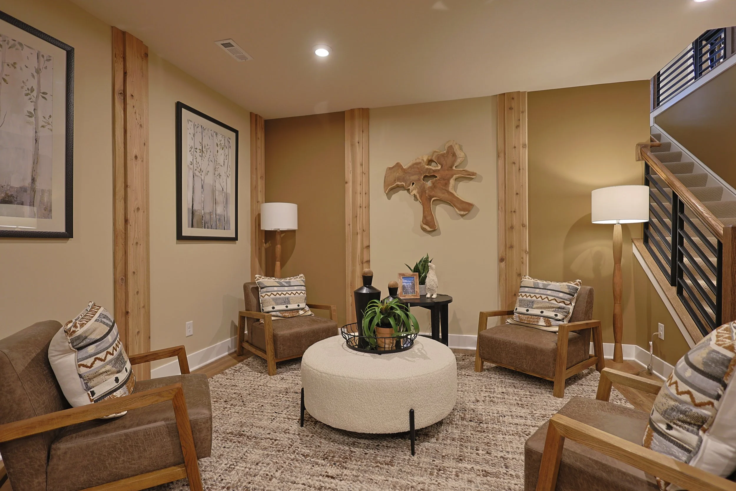Cozy living room with four brown armchairs, a round white ottoman, two floor lamps, framed artwork, wall wood panel accents, and a staircase with black railings.