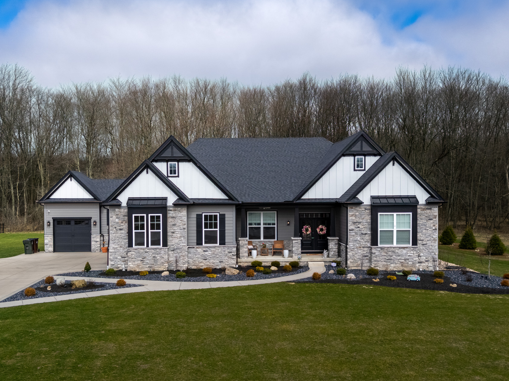 Modern suburban house with stone and siding exterior, gray roof, front porch with two doors decorated with wreaths, well-maintained lawn with flower beds, and a driveway with a garage.