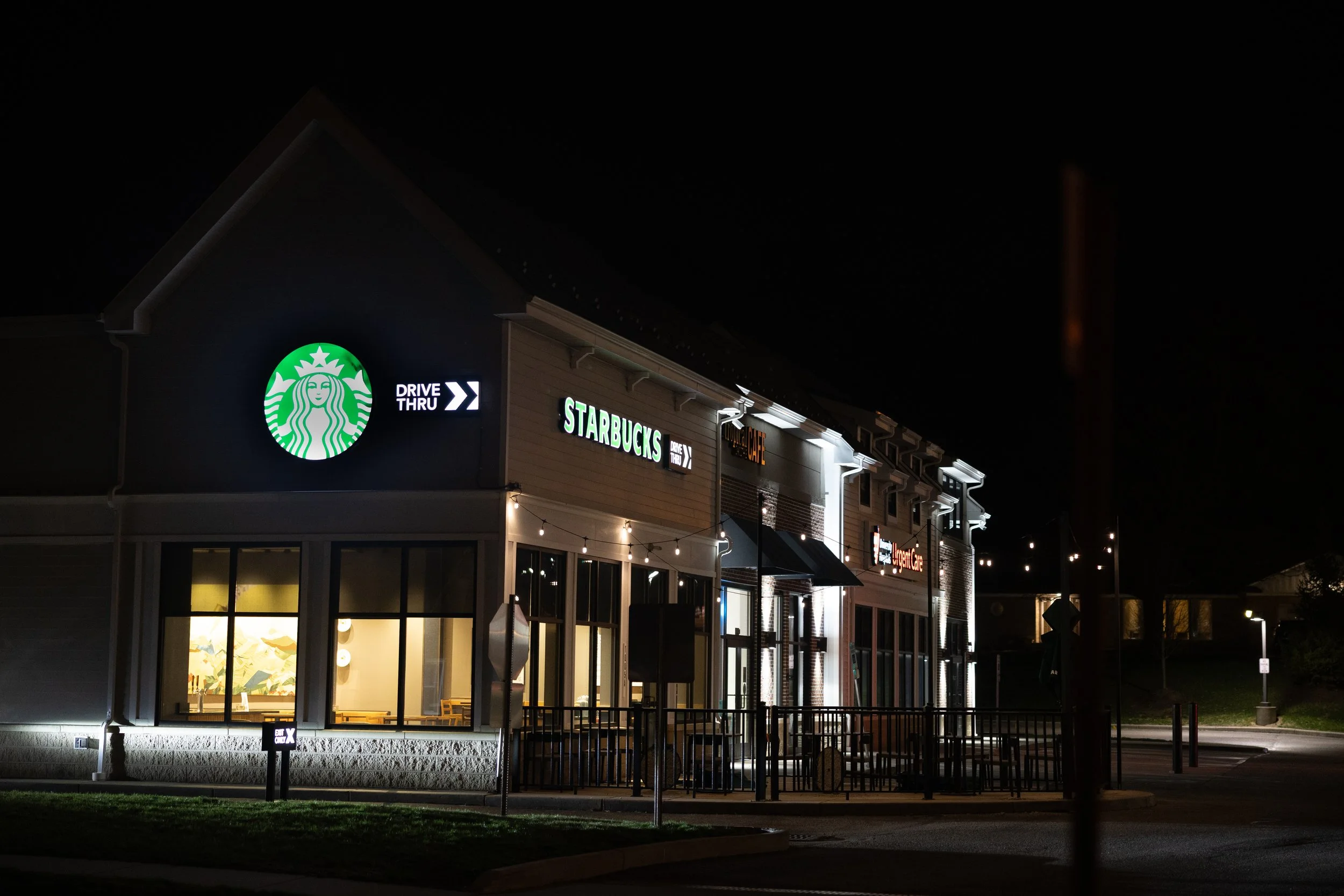 Nighttime view of a commercial building with illuminated signs for Starbucks, including a Drive Thru sign, and other storefronts.