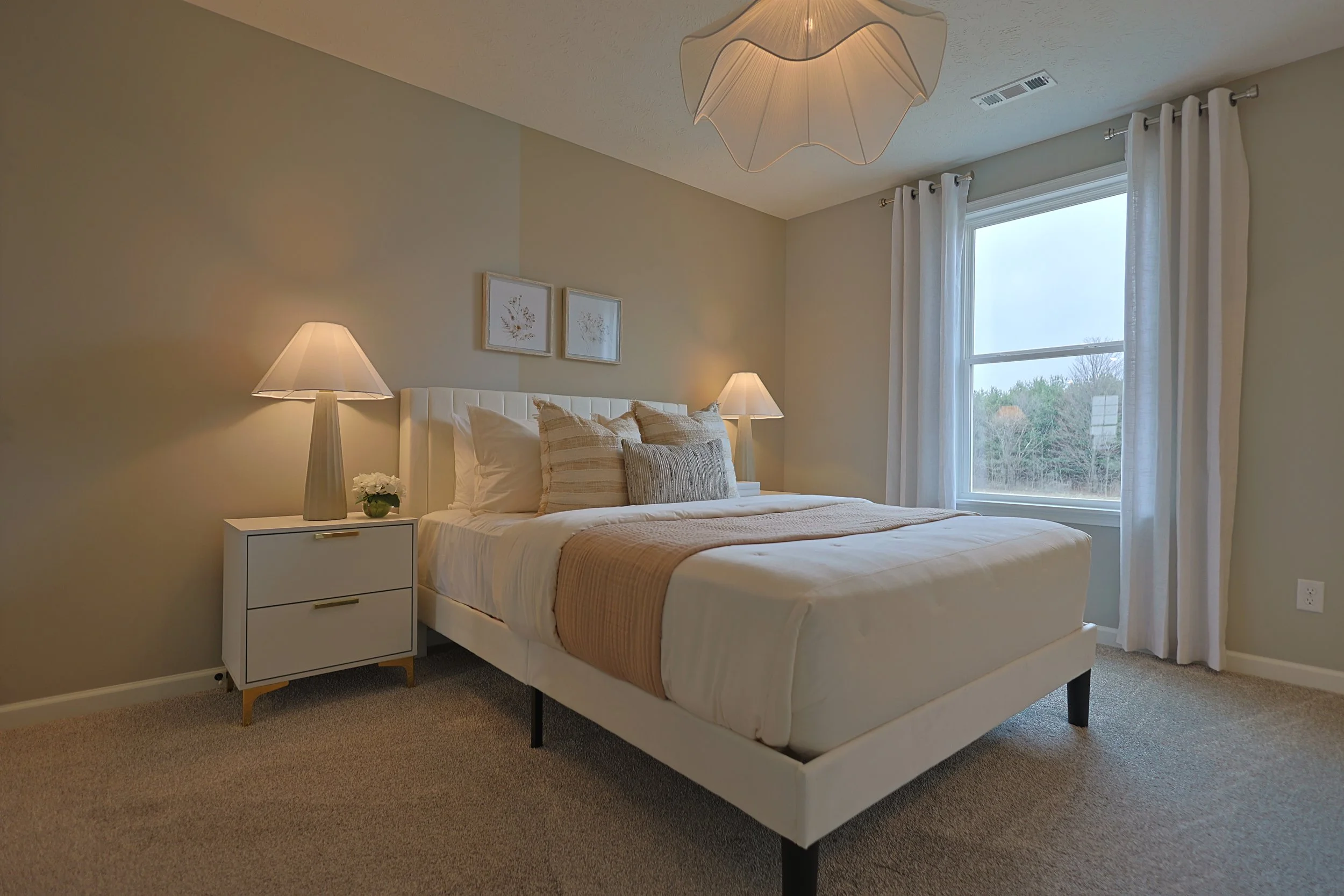 A cozy bedroom with a white bed, beige and cream bedding, two nightstands with lamps, and a window with white curtains showing a view of trees outside.
