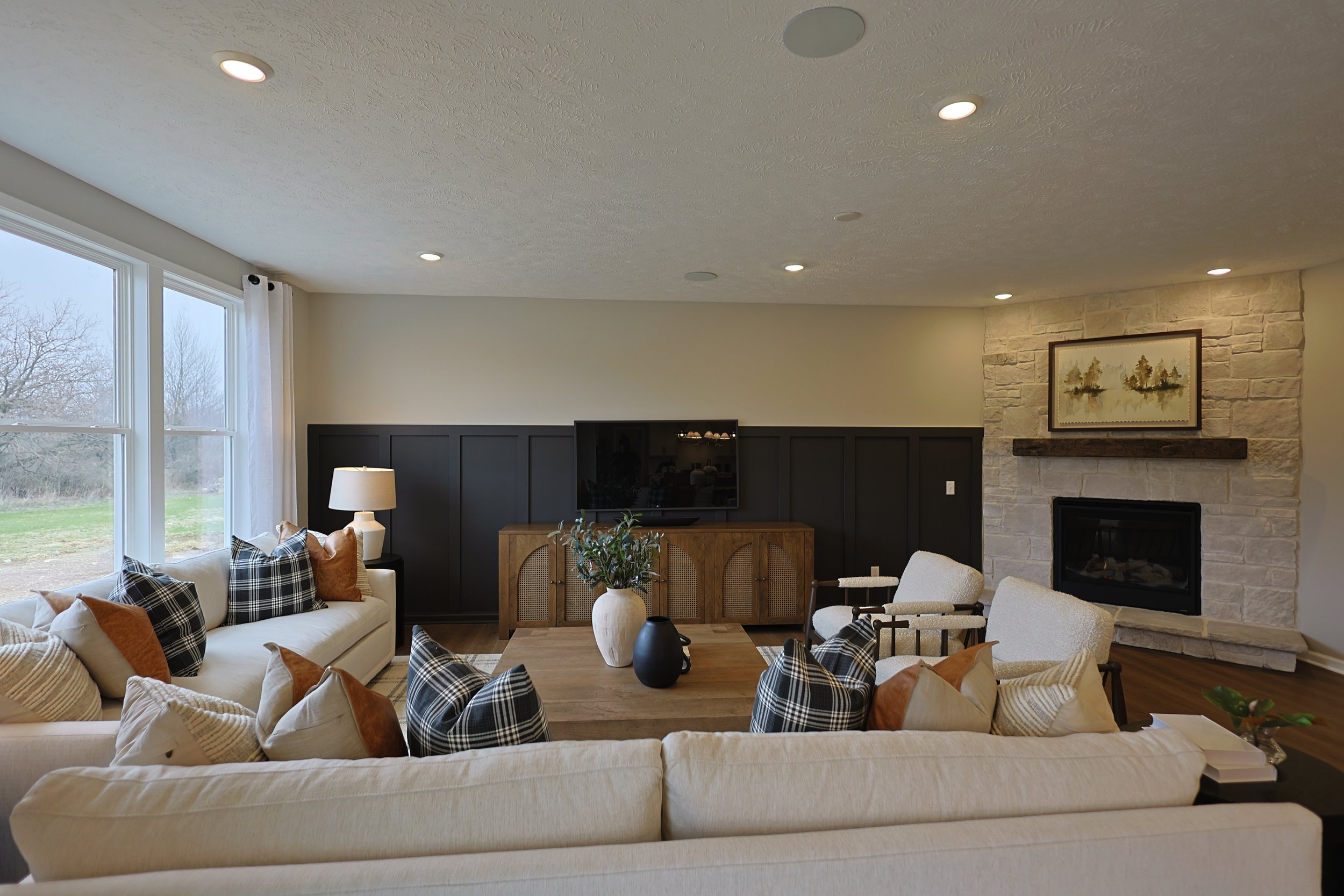 Living room with a large window, beige sofas with plaid and solid pillows, a wooden coffee table with vases, a fireplace with a stone surround, a framed landscape painting, and a TV on a wooden cabinet.