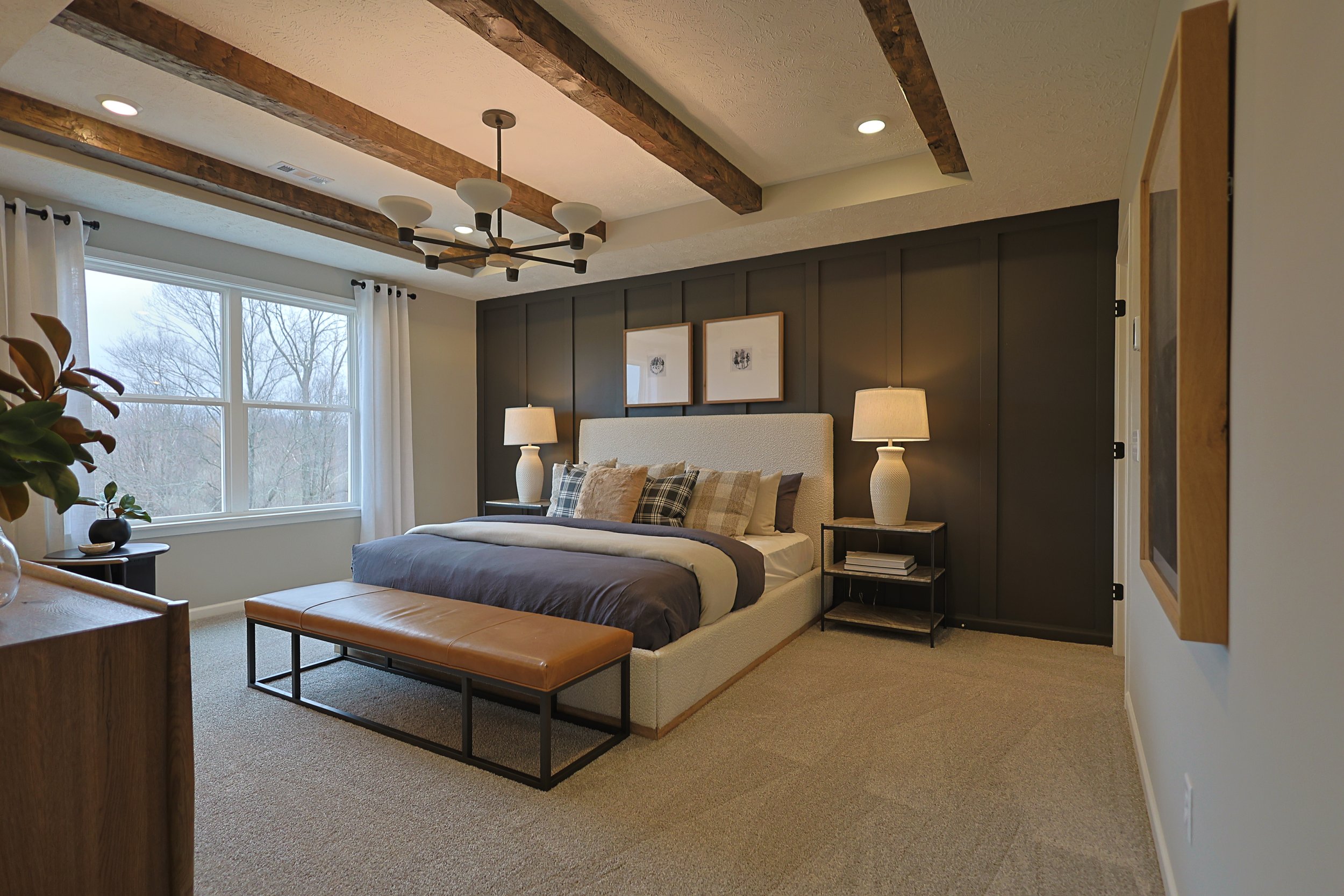 A cozy bedroom with a large window, beige carpeting, and modern decor. There is a bed with multiple pillows, two nightstands with lamps on either side, and a leather bench at the foot of the bed. The ceiling has exposed wooden beams and a stylish cha