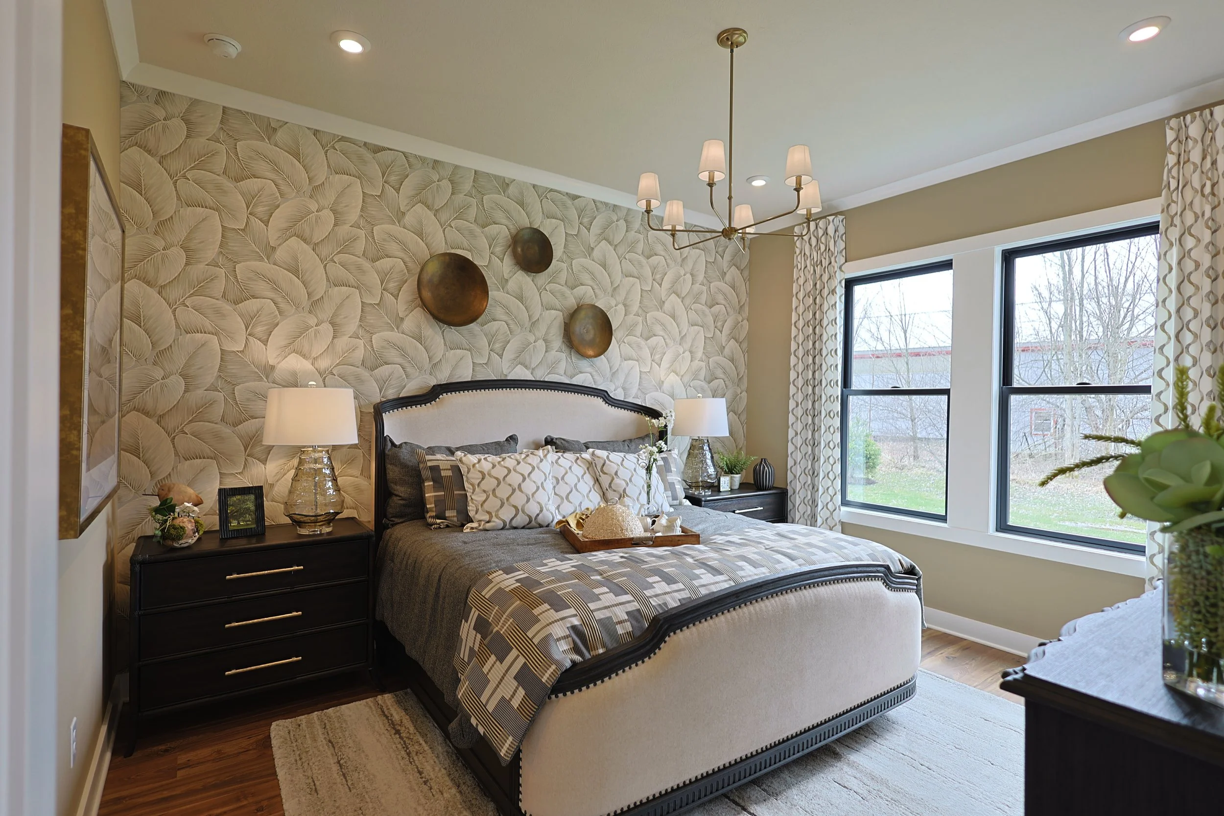 A bedroom with a decorated wall behind a bed, two nightstands with lamps, large windows, and curtains, with some plants and decorative items.
