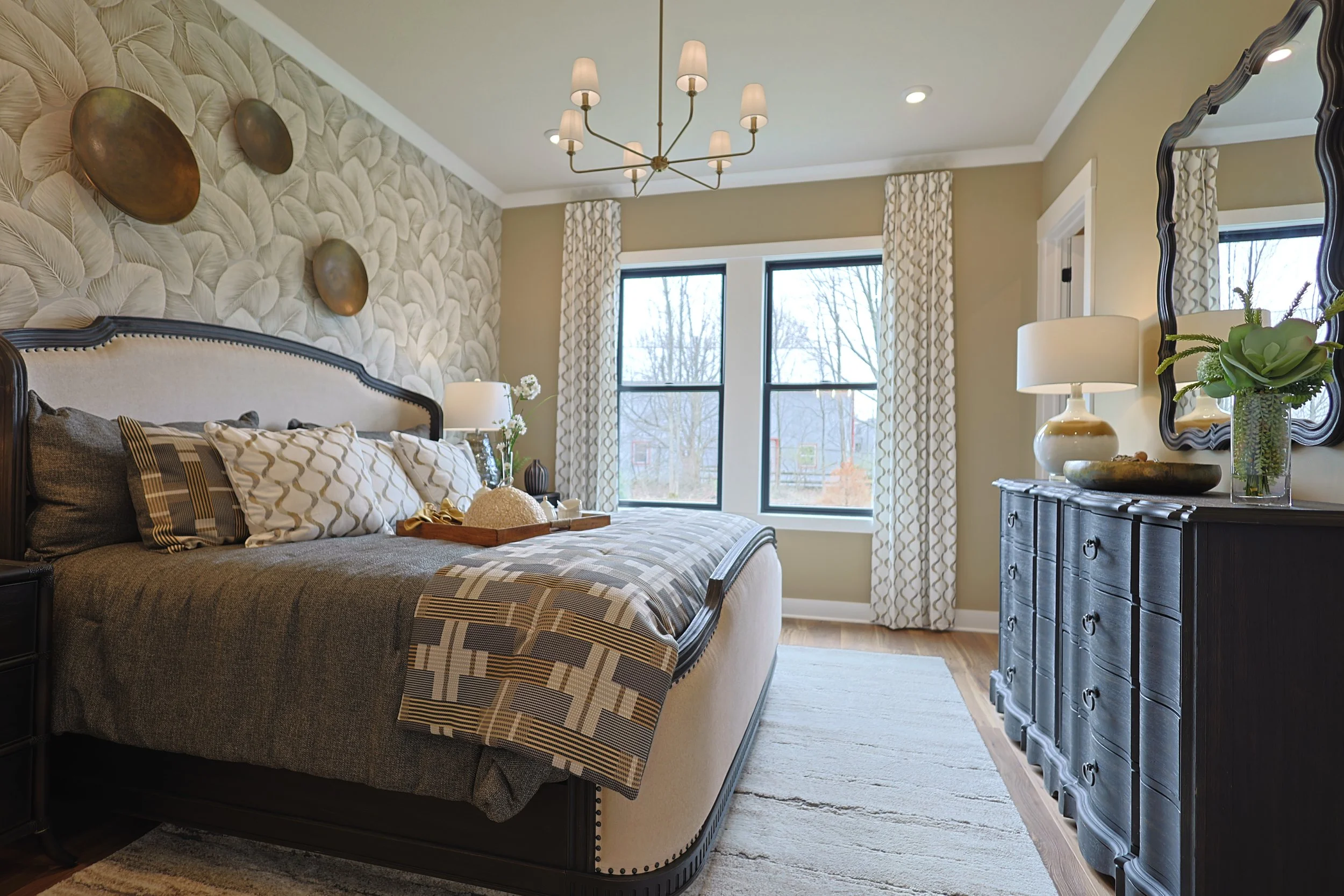 A bedroom with a large bed, dark wooden furniture, beige and patterned pillows, a textured wall with decorative wall art, a window with patterned curtains, and a mirror on the wall.