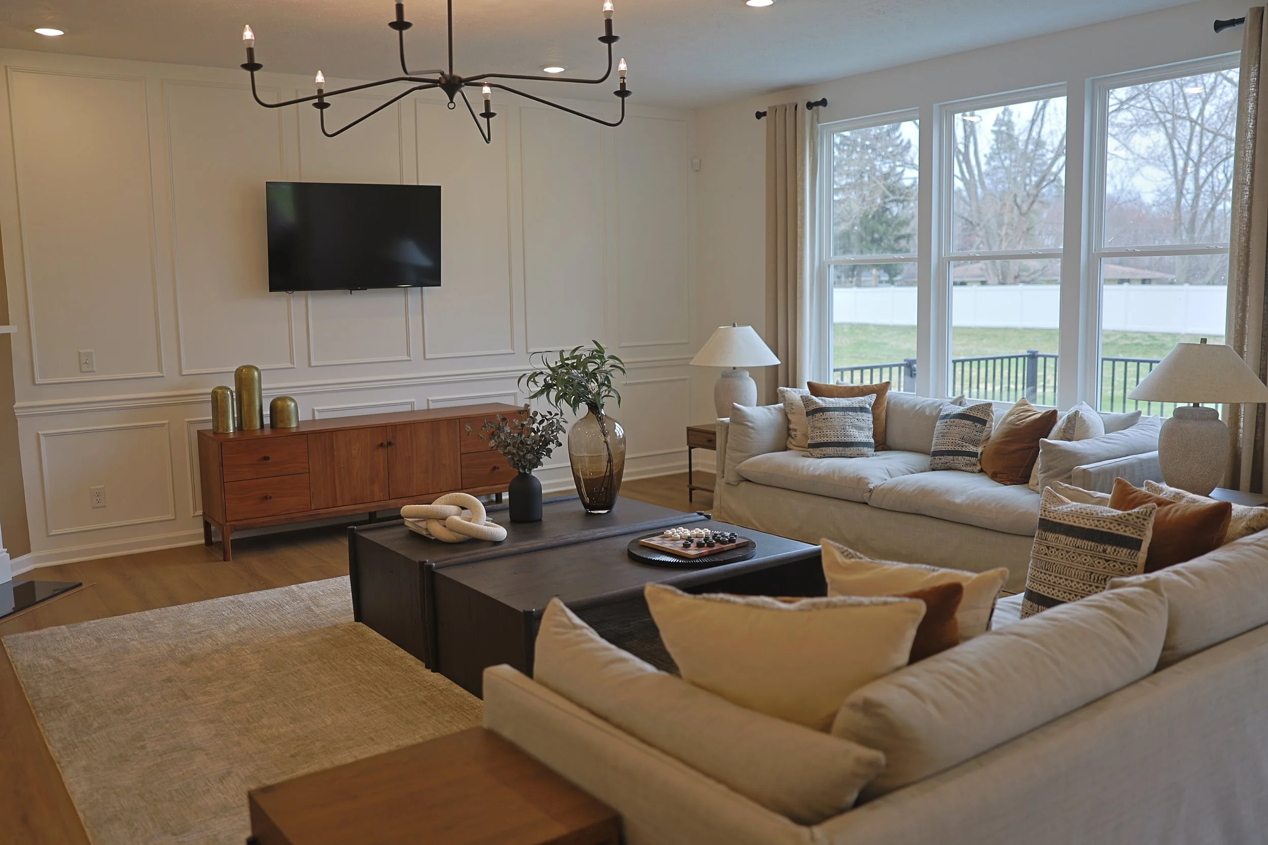 Living room with white sofas, wooden coffee table, beige and patterned pillows, side tables with lamps, large windows with beige curtains, wall-mounted TV, wooden console with decorative vases, and a chandelier.
