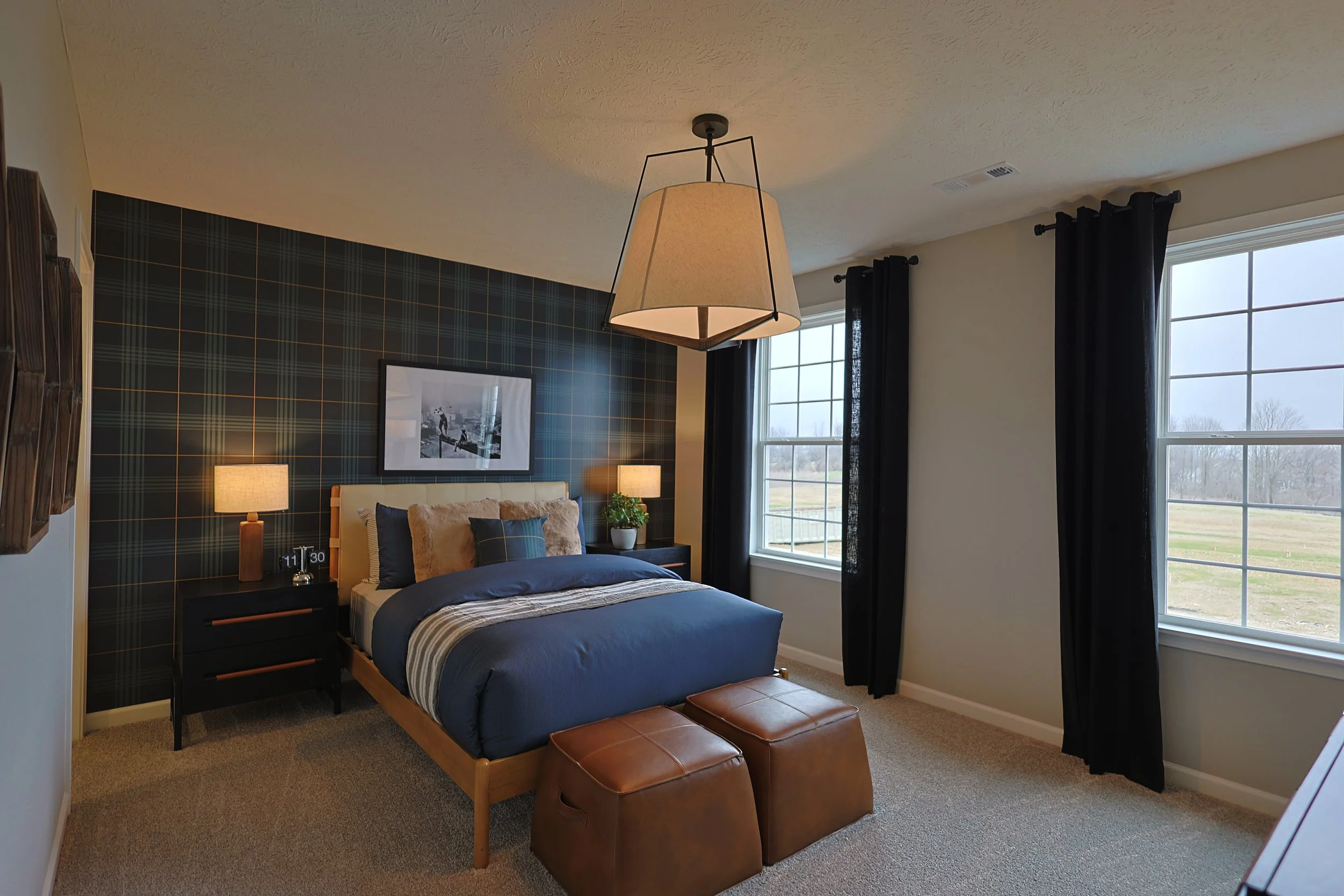A bedroom with a bed having navy blue and gray bedding, two bedside tables with lamps, a plaid accent wall behind the bed, large windows with black curtains, and two brown leather ottomans at the foot of the bed.