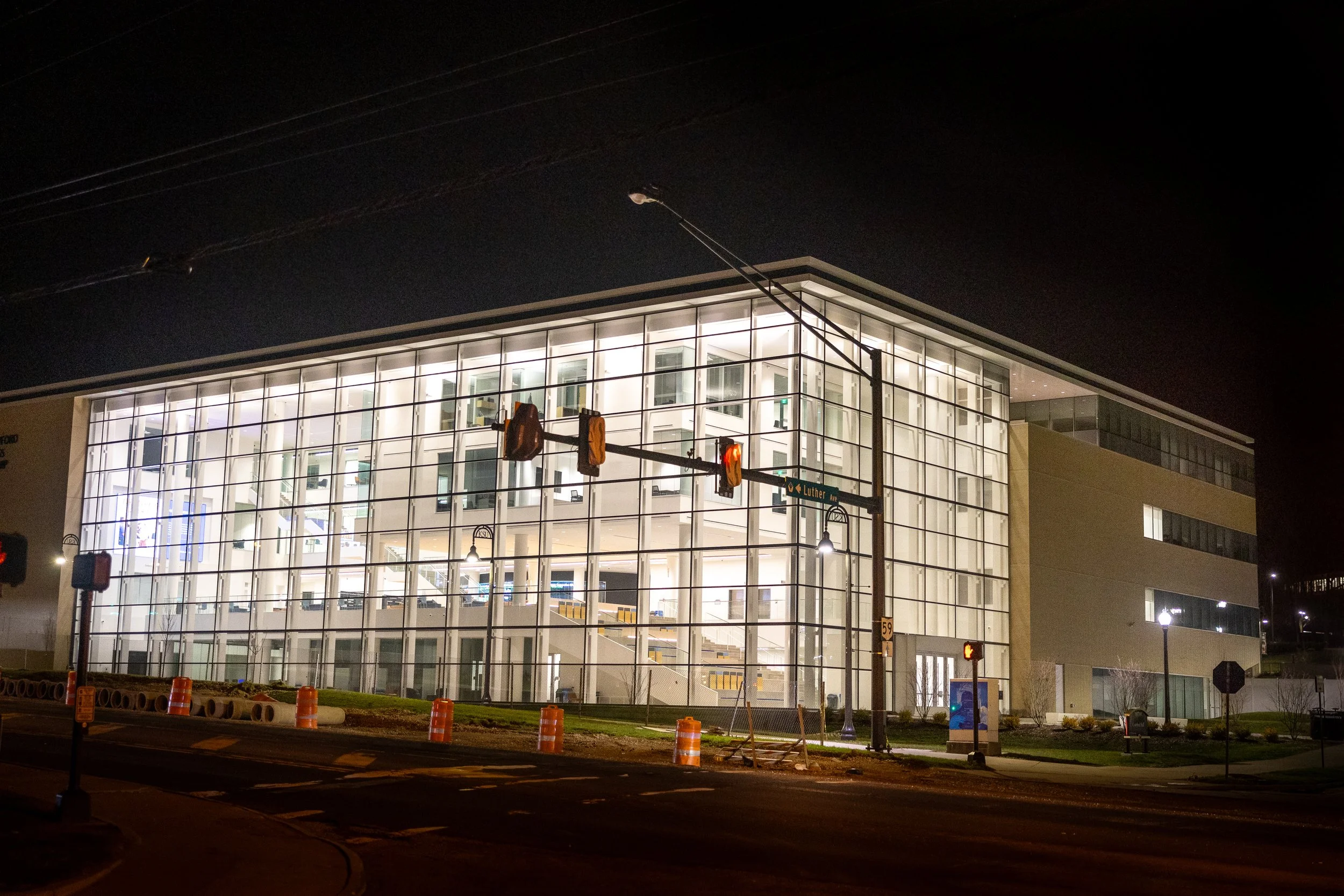 Modern multi-story building with large glass windows illuminated at night, with street traffic lights and street signs visible in the foreground.
