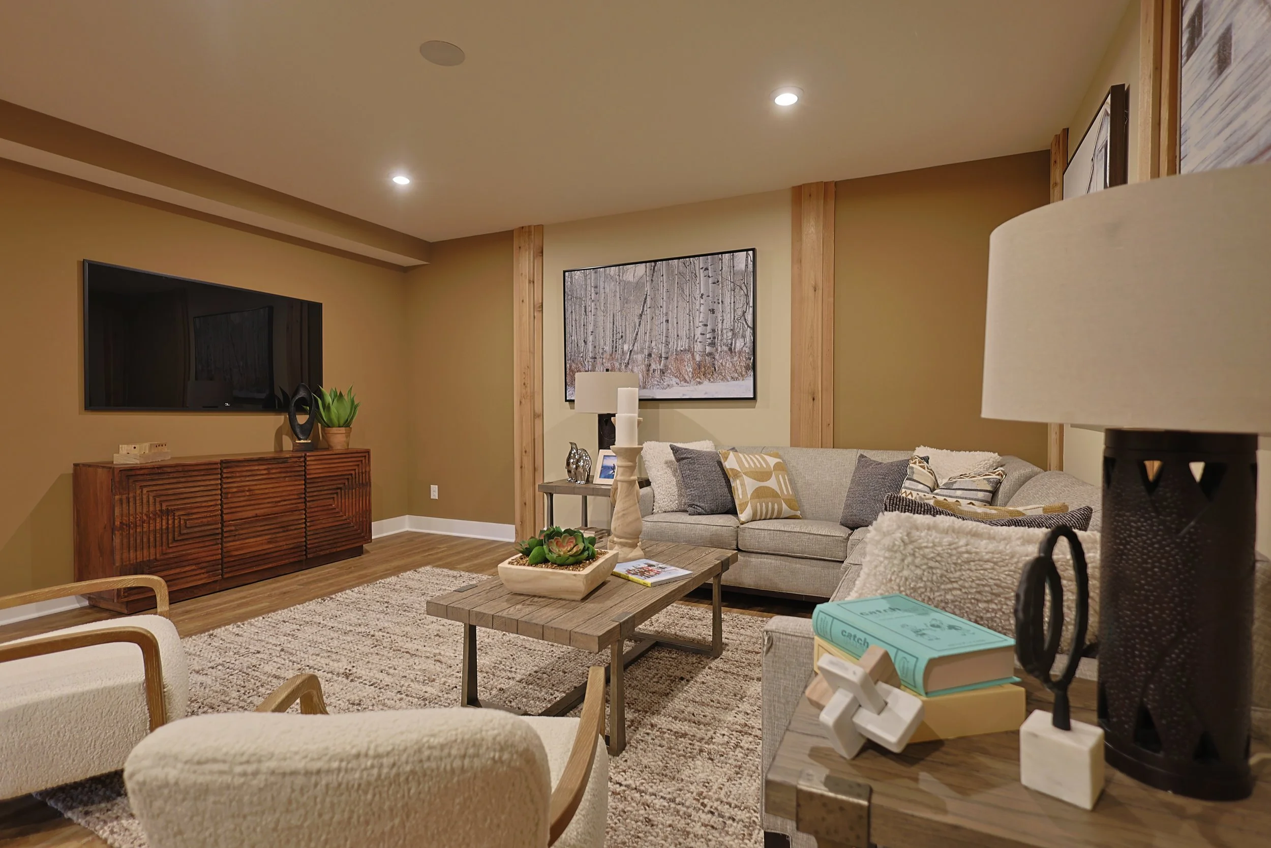 Living room with beige walls, hardwood floors, and modern decor, including a gray sofa with patterned pillows, a wooden coffee table, a TV on a wooden console, and various decorative items.