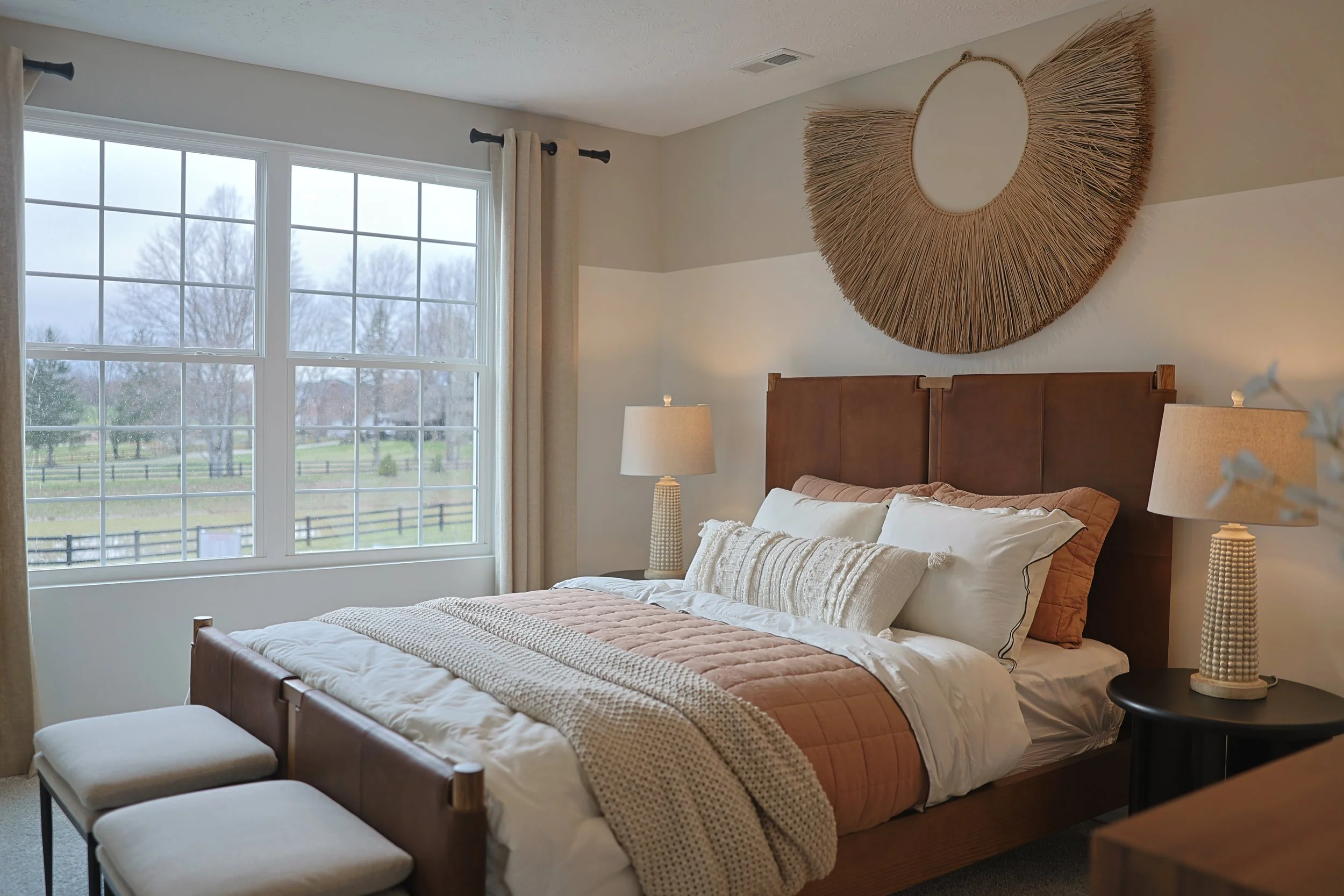 Cozy bedroom with large window, beige curtains, wooden bed with white and beige bedding, wicker wall art above headboard, two table lamps with textured bases on black side tables.