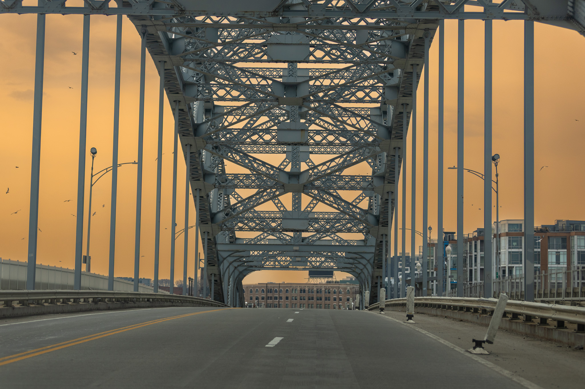 A view underneath a metal bridge with the road leading to a cityscape at sunset.
