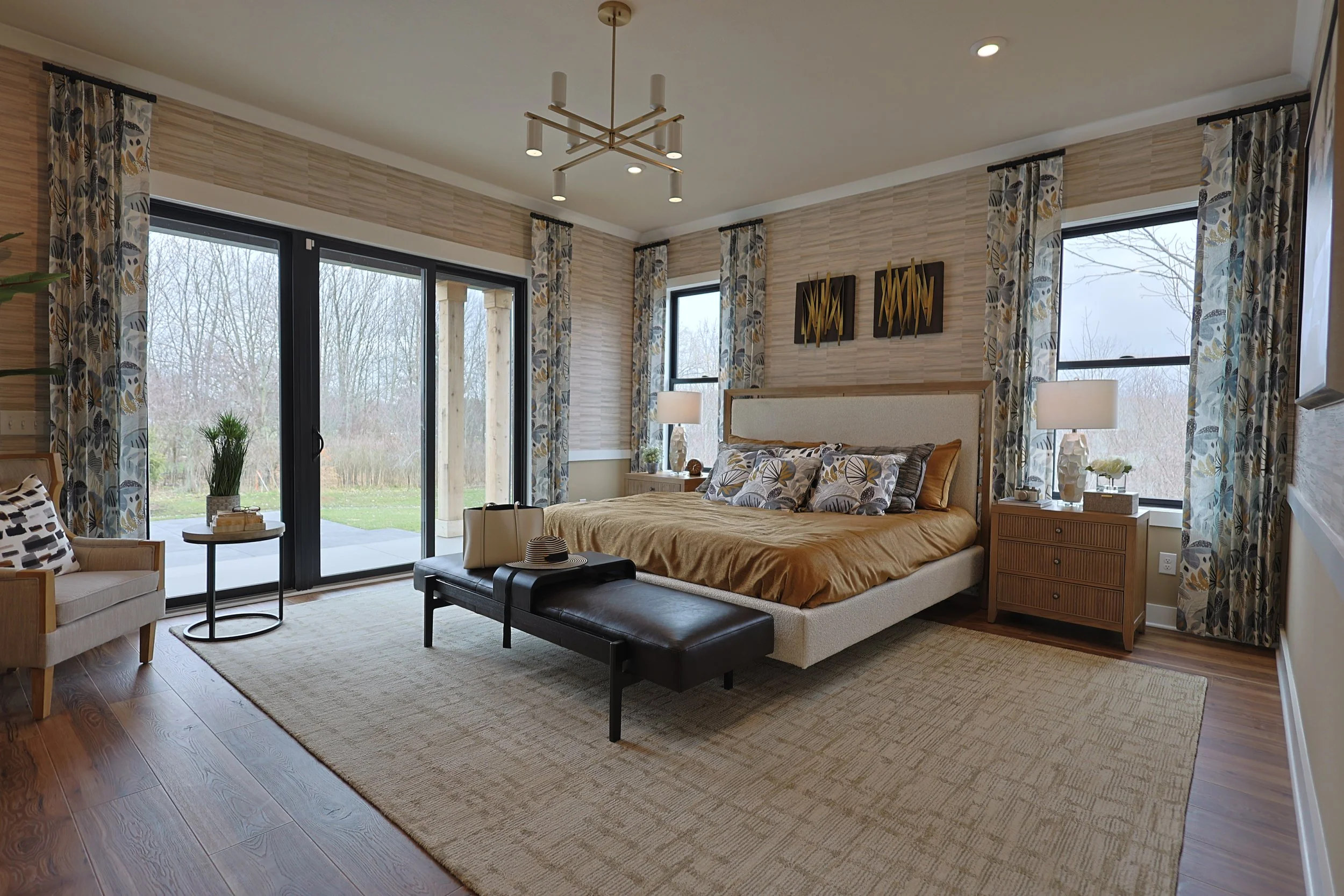 A modern bedroom with a beige upholstered bed, patterned pillows, and two nightstands with lamps, next to large windows with floral curtains and a sliding glass door leading outside.