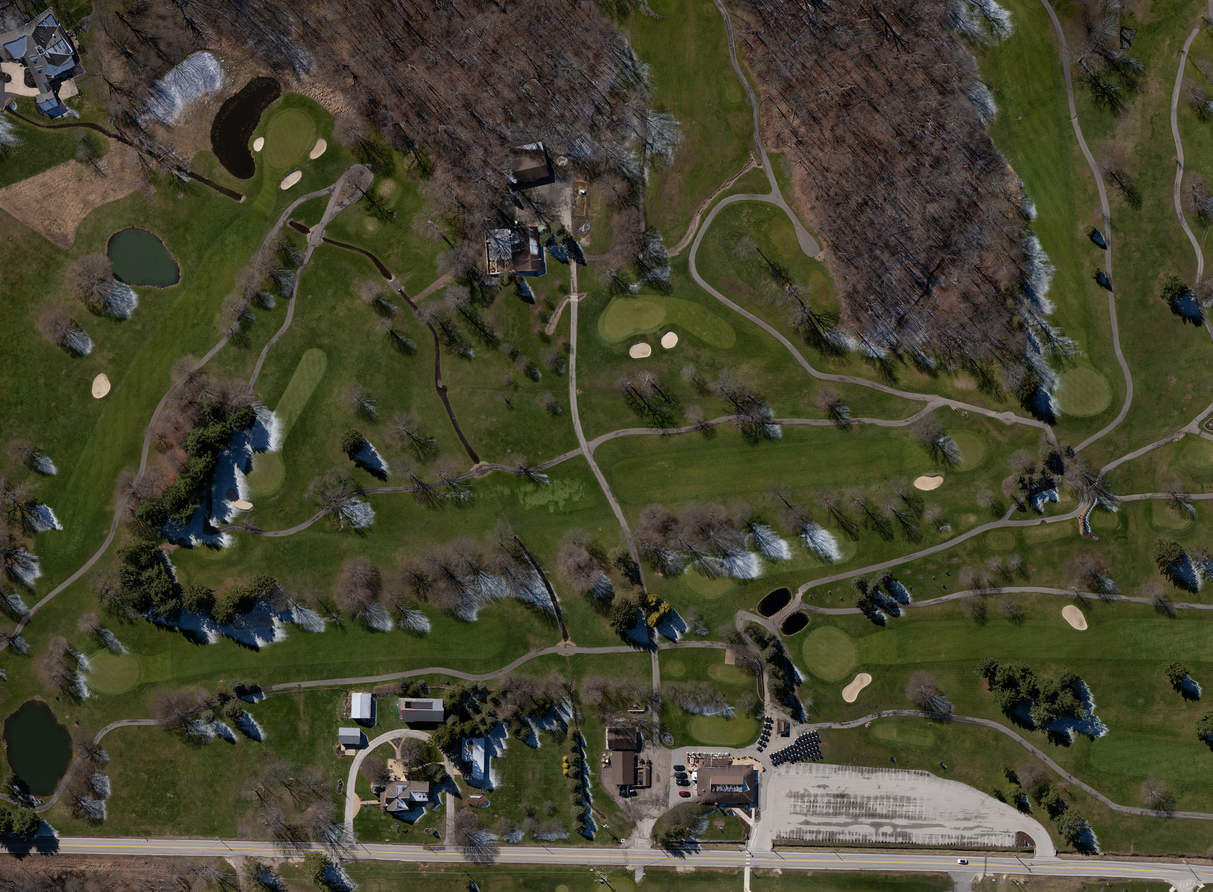 Aerial view of a golf course with multiple fairways, putting greens, water hazards, trees, and a clubhouse area with parking lot.