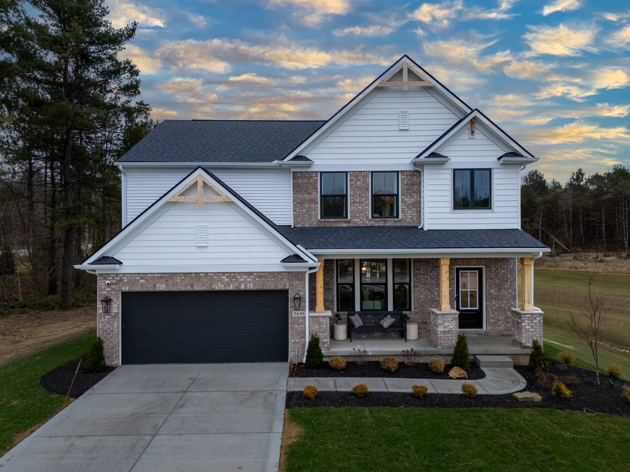 Front view of a new, two-story house with a brick and white siding exterior, black garage door, and a small front porch with a bench and pillows, surrounded by newly planted bushes and a landscaped yard, under a partly cloudy sky at sunset.