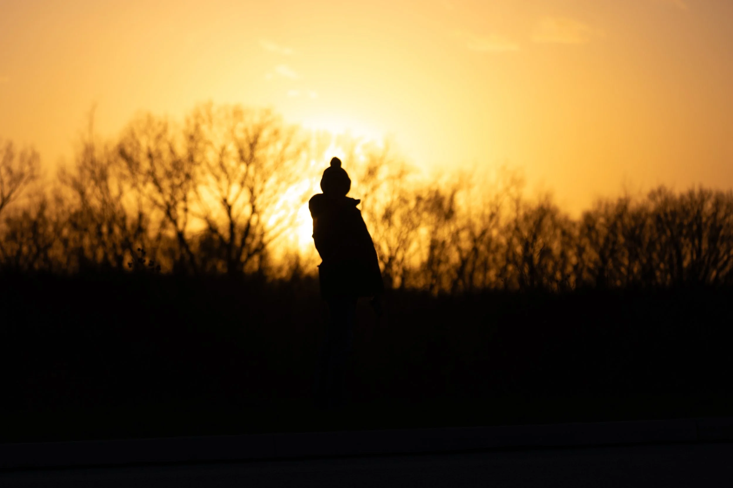 Silhouette of a person with a hat walking outdoors during sunset with trees in the background.