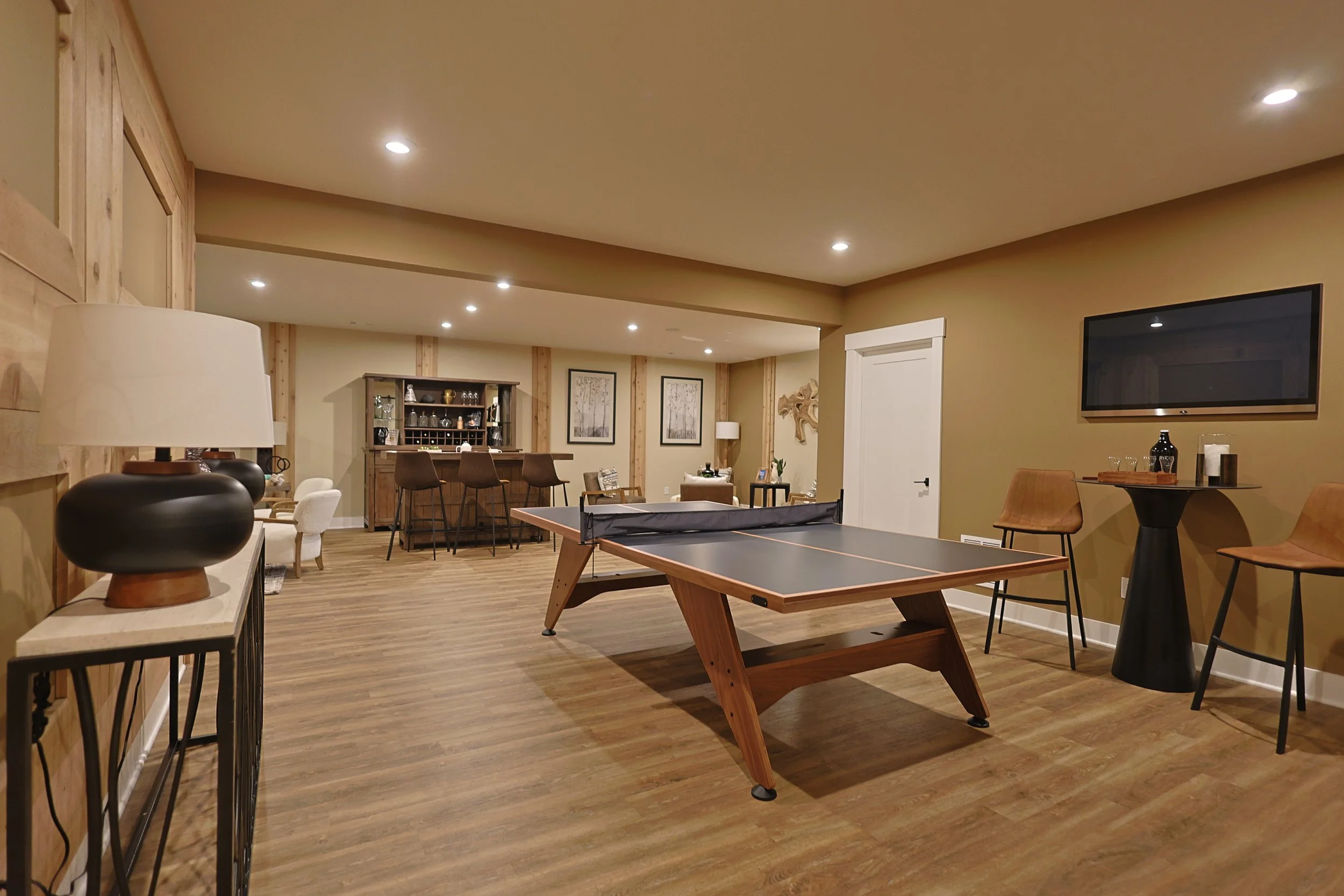 A spacious basement recreation room with a ping pong table, bar area, seating, and a mounted flat-screen TV on a tan wall.