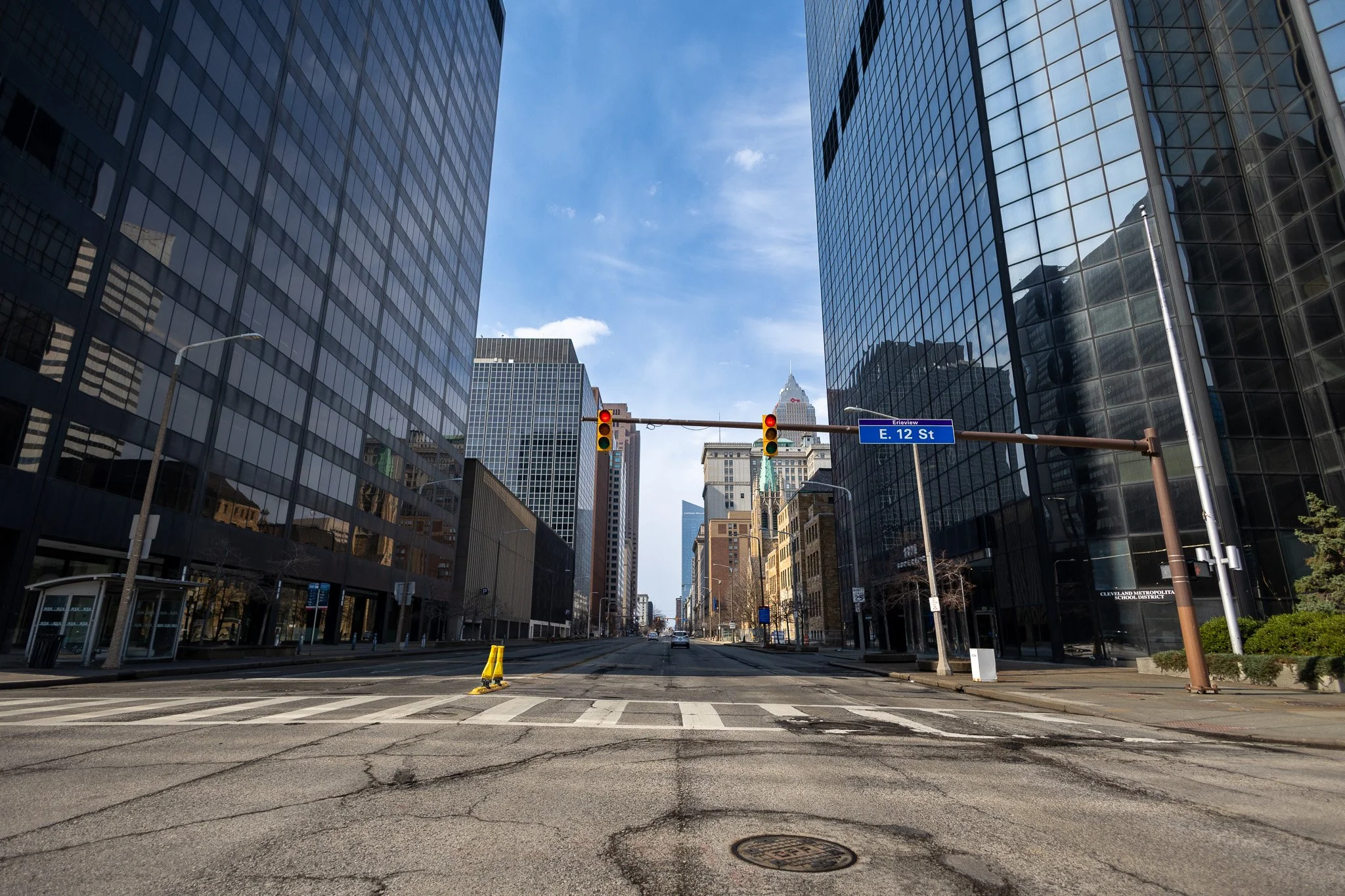 Empty city street with tall glass buildings on both sides, traffic lights, and a blue street sign reading 'E. 12 St' under a partly cloudy sky.