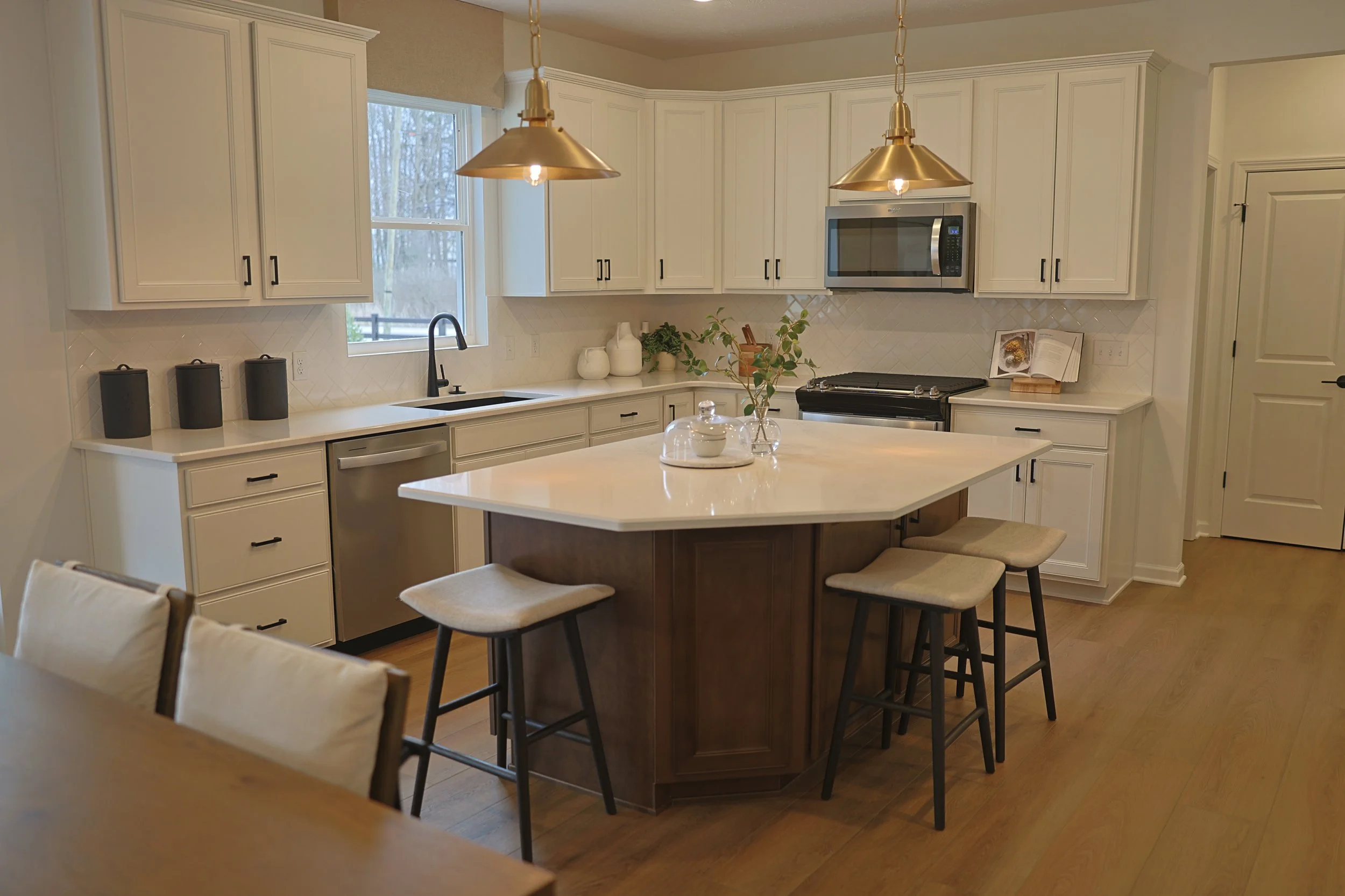 Modern kitchen with white cabinets, a kitchen island with a dark base, beige stools, and stainless steel appliances. Open cookbook and small decorative items on counter, pendant lights above island.