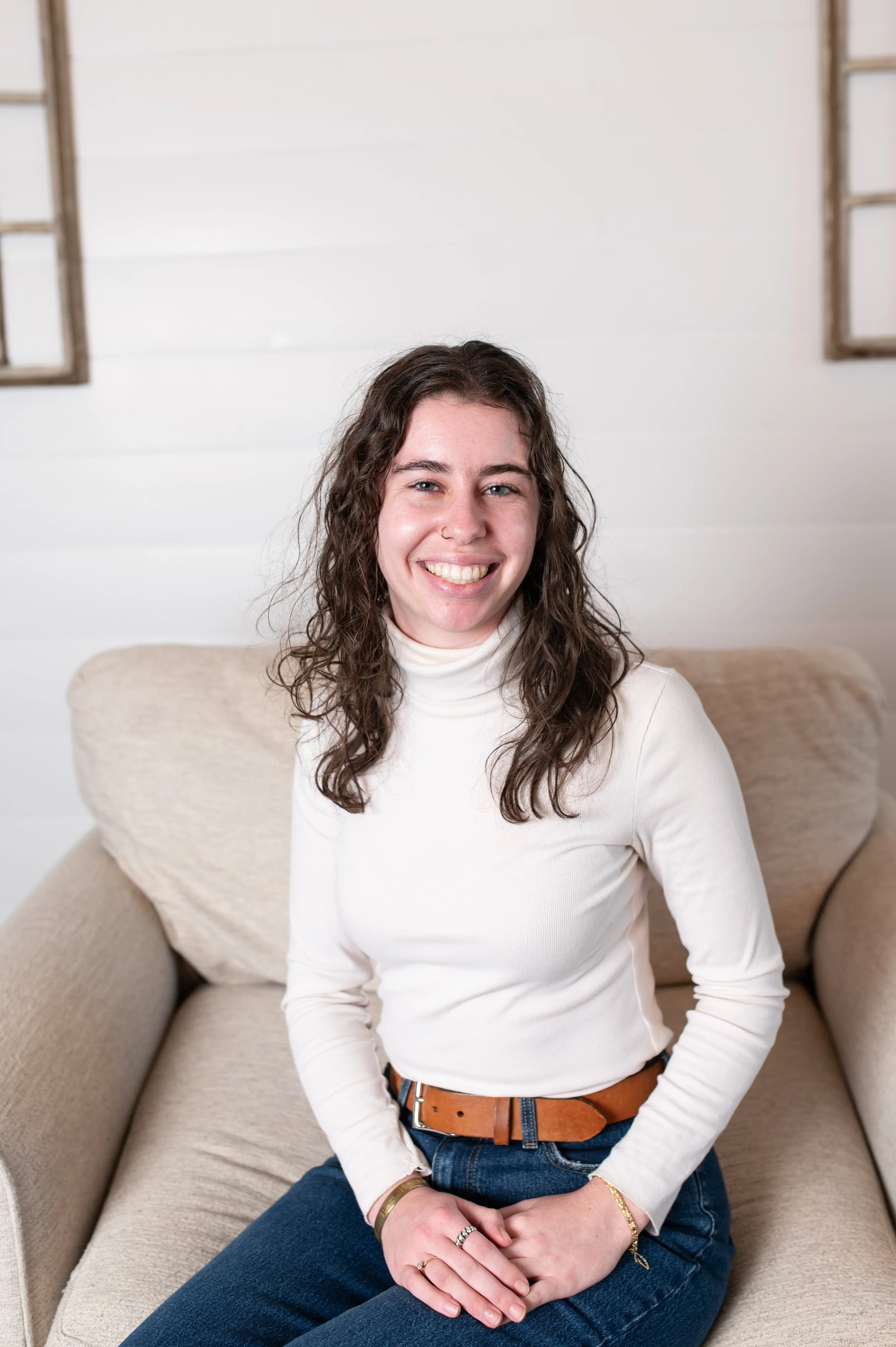 Retrato de una joven sonriendo, sentada en un sillón beige, con fondo de pared de tablas blancas y ventanas de madera a cada lado.