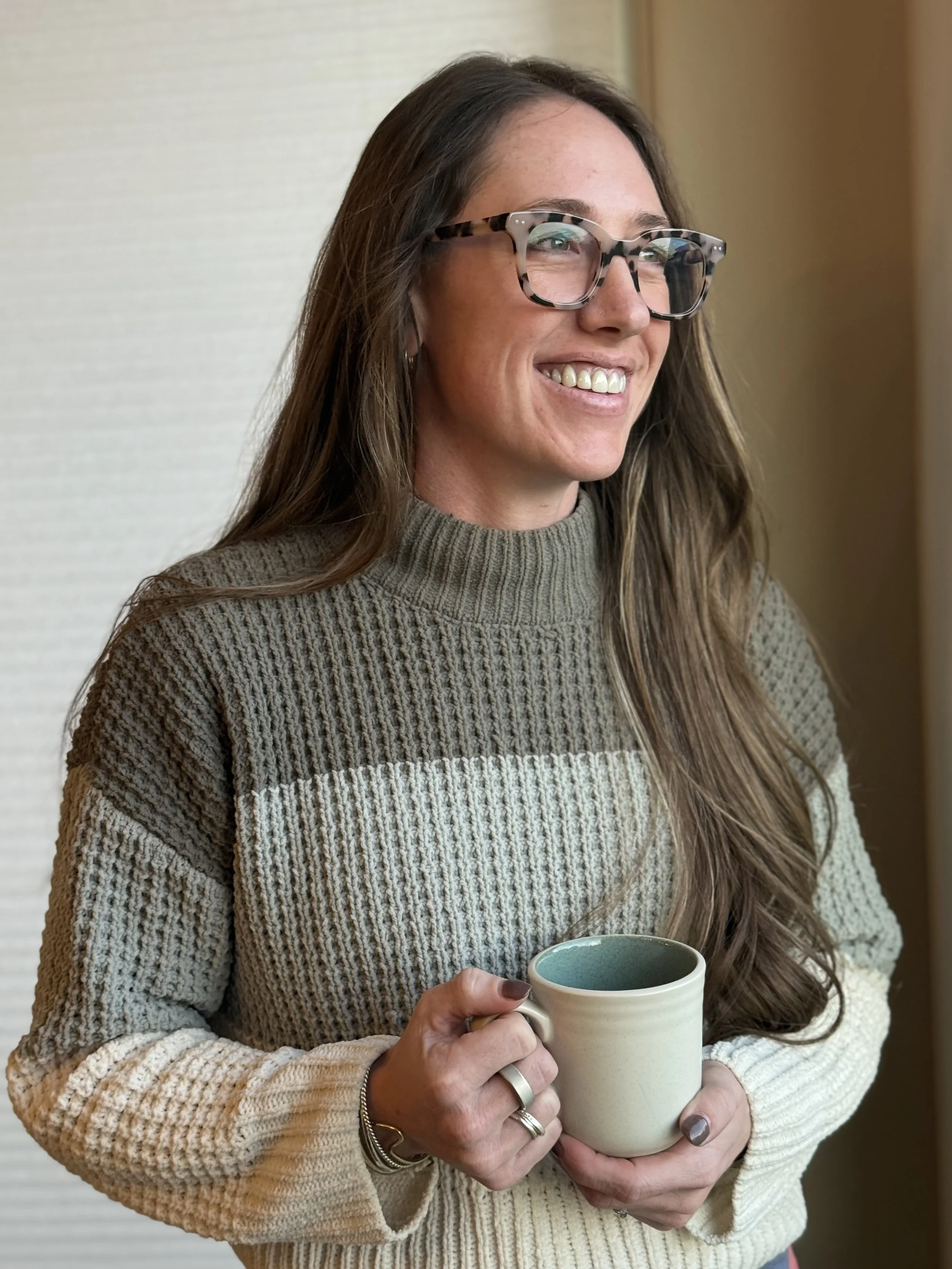 A woman with long hair and glasses smiling while holding a mug, wearing a knit sweater.