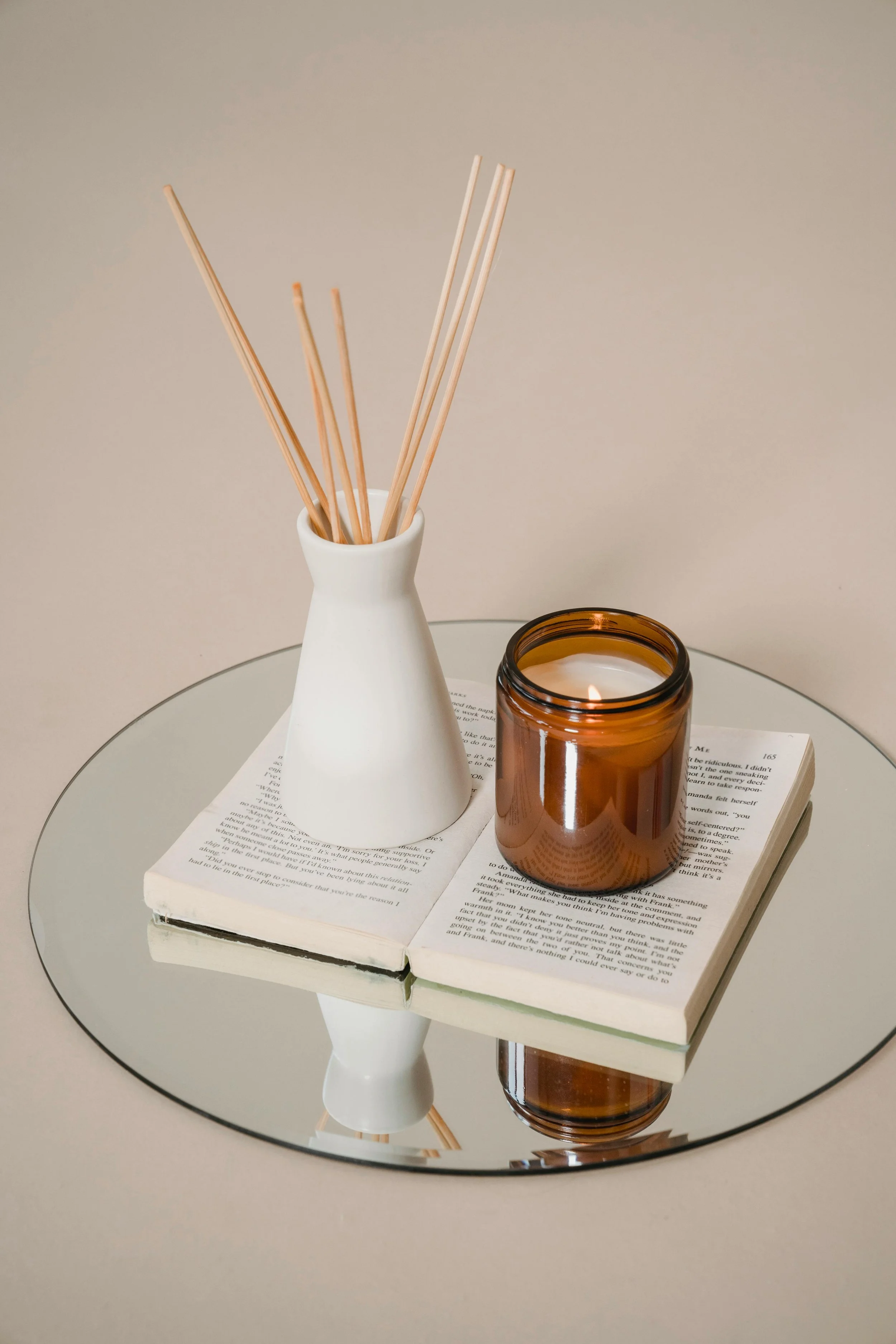 A white vase with wooden stir sticks, a brown candle jar, and an open book reflected on a round mirror surface.