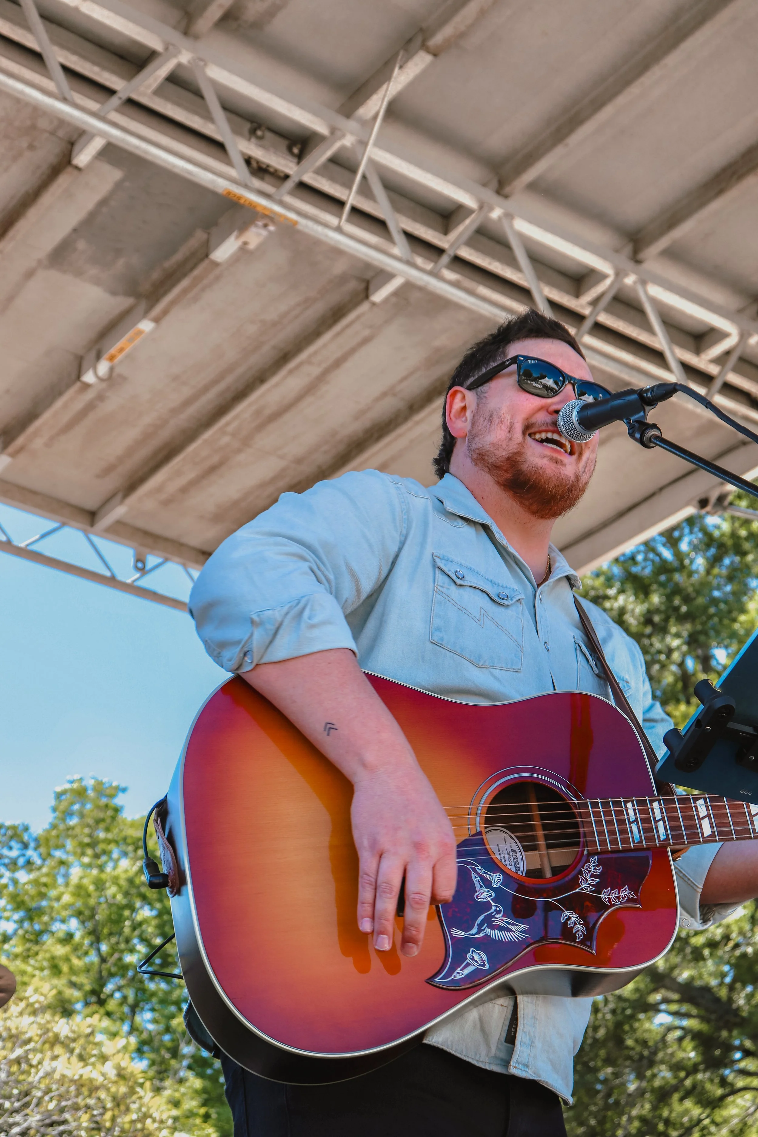 A man wearing sunglasses and a light blue shirt playing an acoustic guitar with decorative art on it, singing into a microphone under a bridge with a cloudy sky in the background.