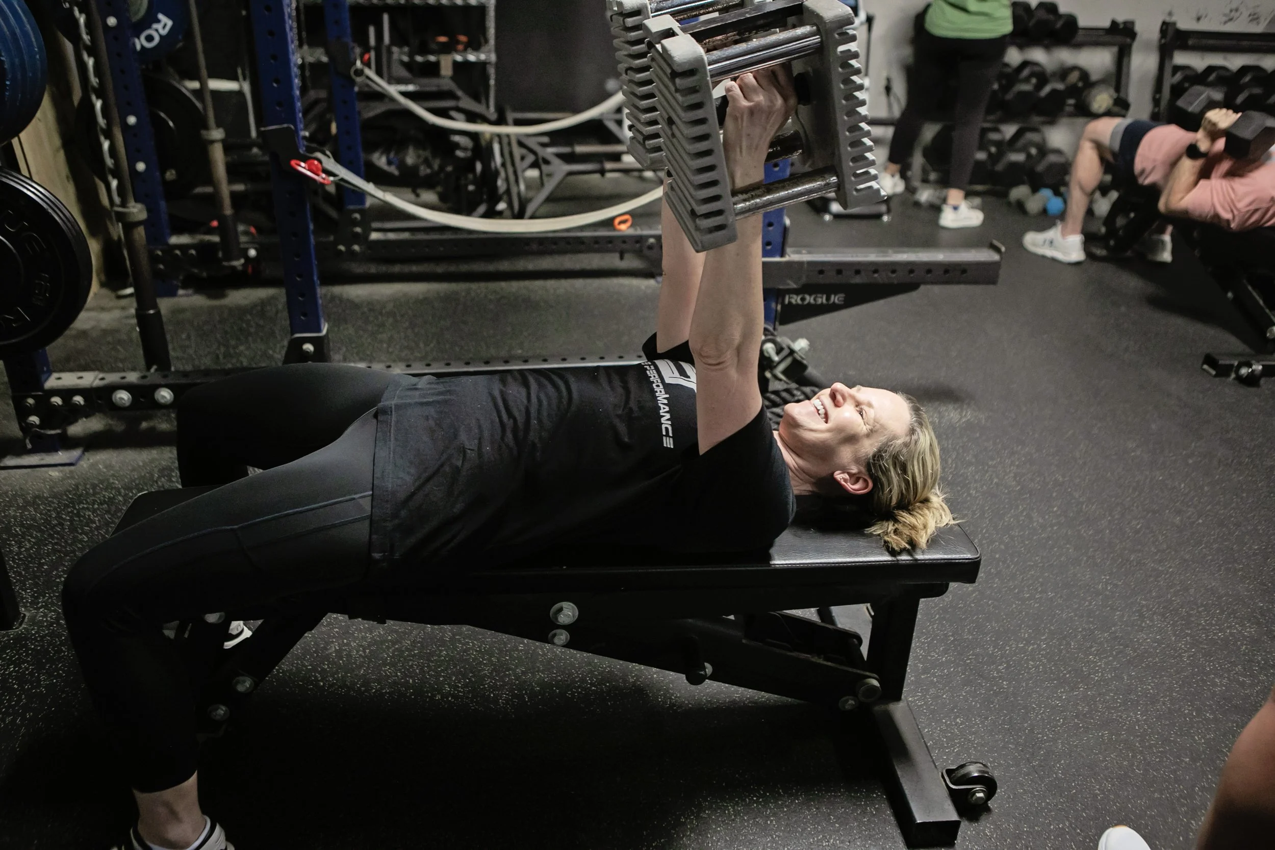 A woman lying on a bench press bench with a dumbbell in each hand, smiling during a workout at Edge Performance in Valdosta, GA