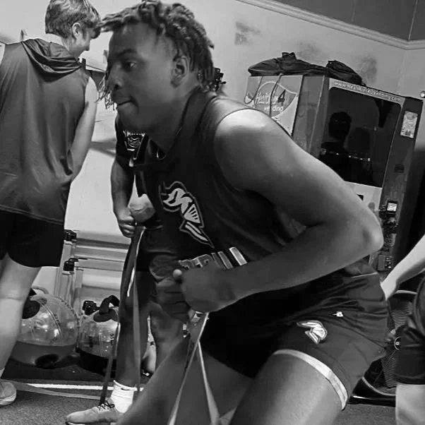 A young man in athletic shorts and a sleeveless shirt doing resistance training with a resistance band in a gym, while other people are exercising in the background.