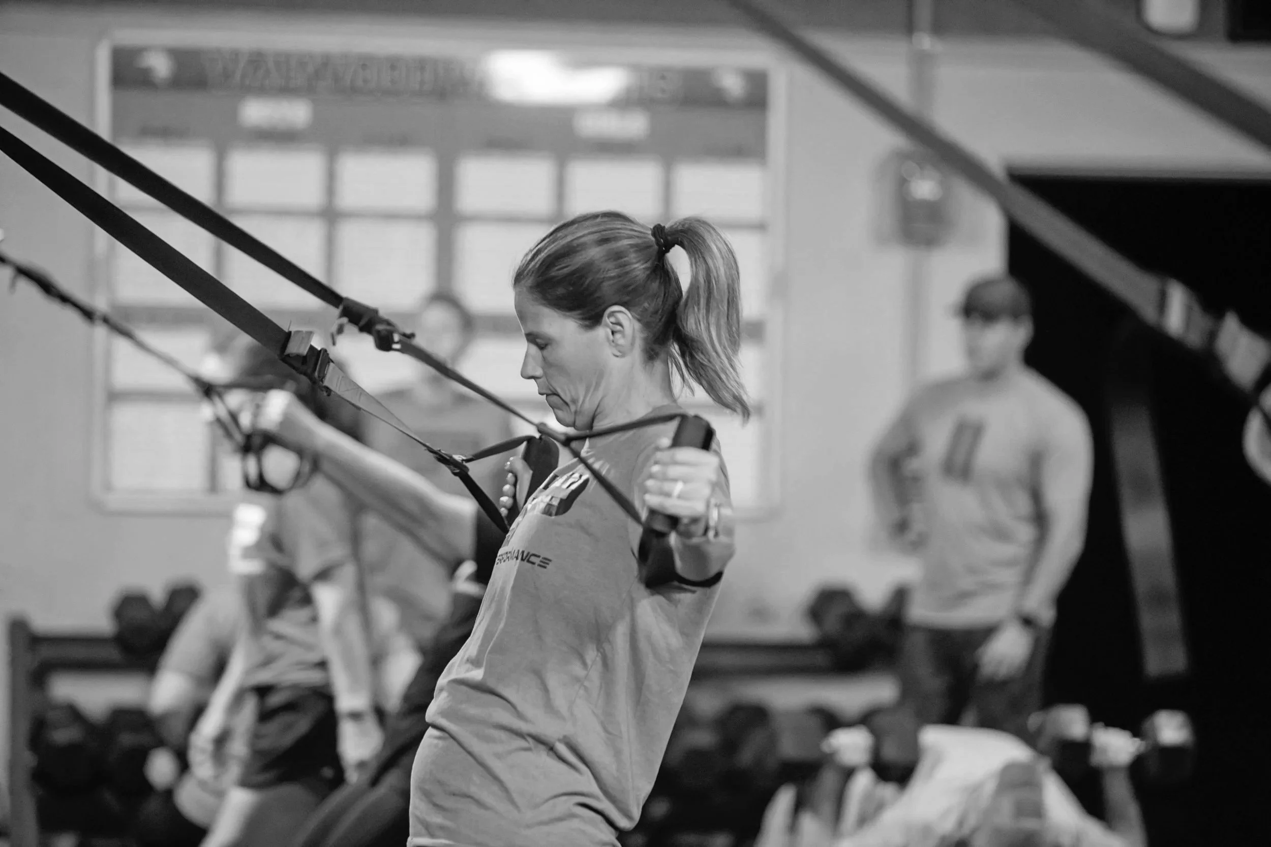 Woman working out with TRX suspension straps in a gym, with other people exercising in the background.