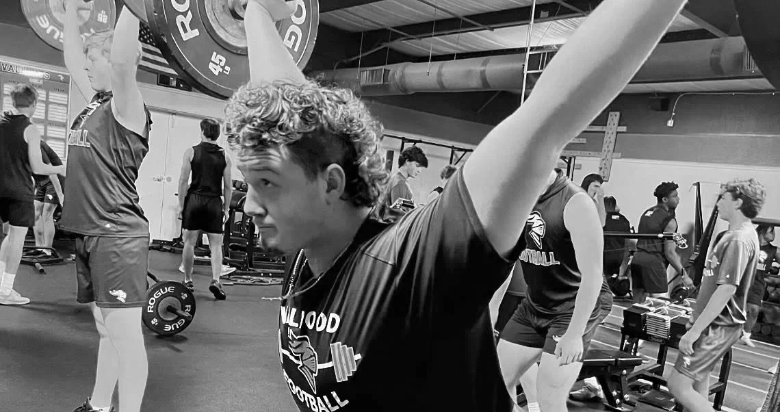 Young man with curly hair lifting a barbell with weights in a gym, surrounded by other athletes training.