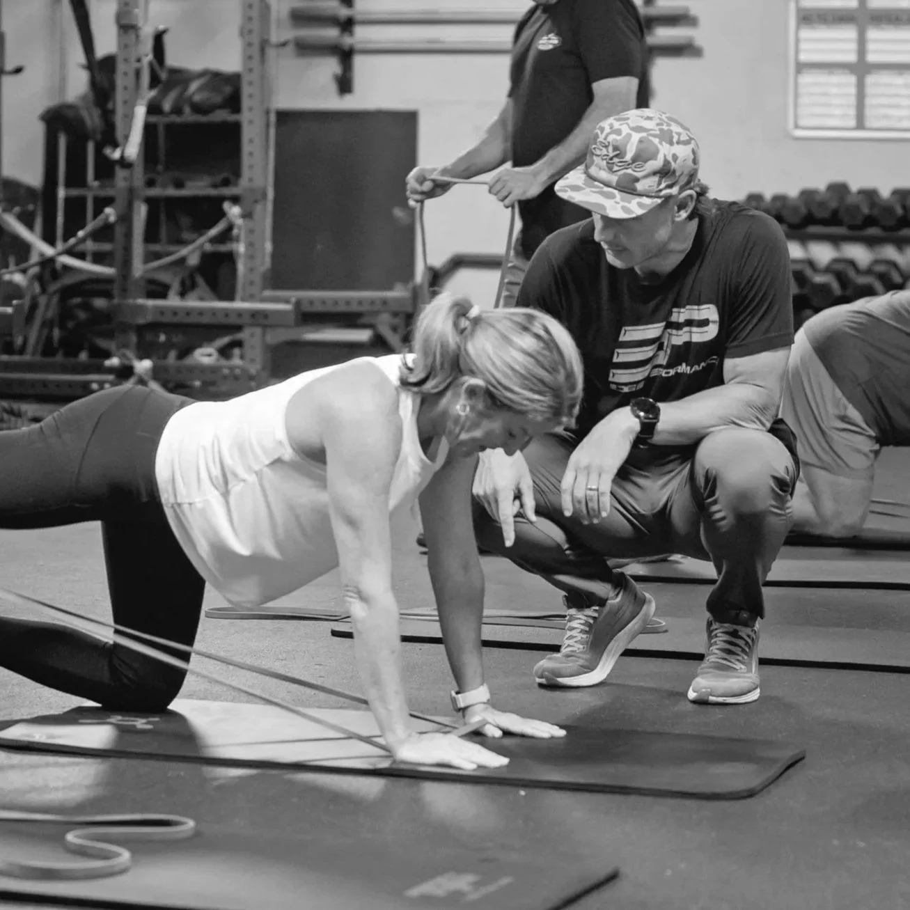 A woman is doing an exercise with resistance bands in a gym, being guided by a trainer, while another person exercises in the background.