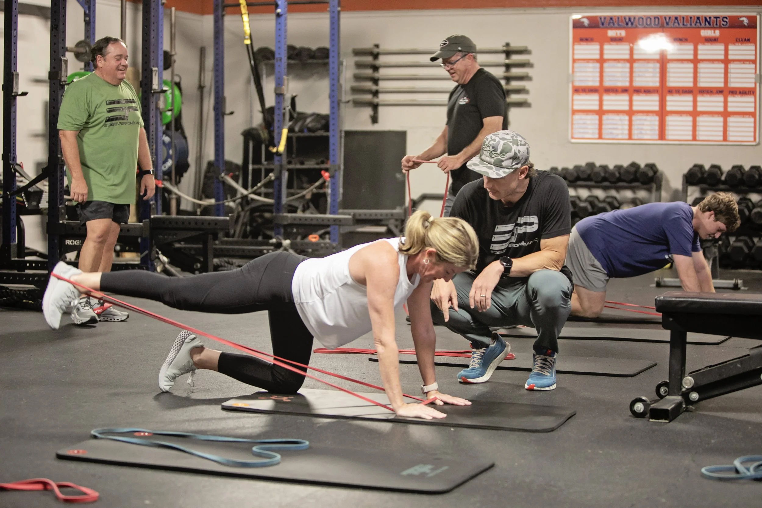 A group workout session with a woman doing resistance band exercises on a mat at a gym, surrounded by men observing and assisting, with gym equipment and a motivational chart in the background.