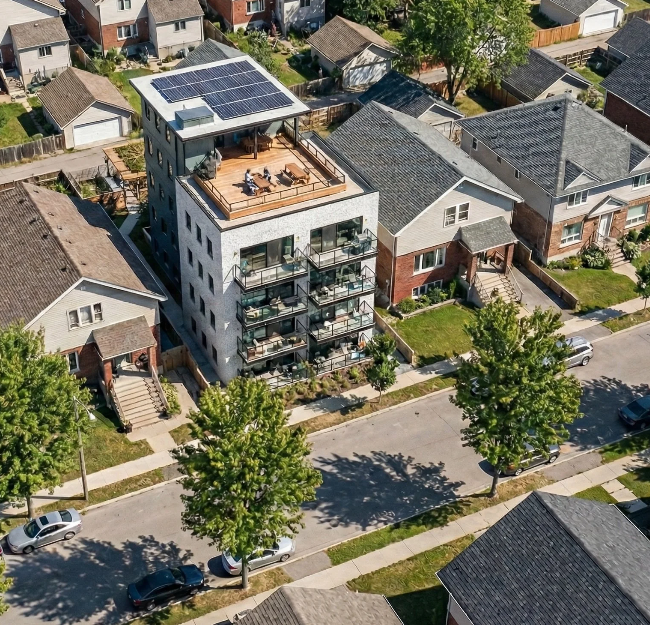 A multi-story residential building with solar panels on the roof and a rooftop deck featuring outdoor furniture, surrounded by nearby houses and trees in a neighborhood.