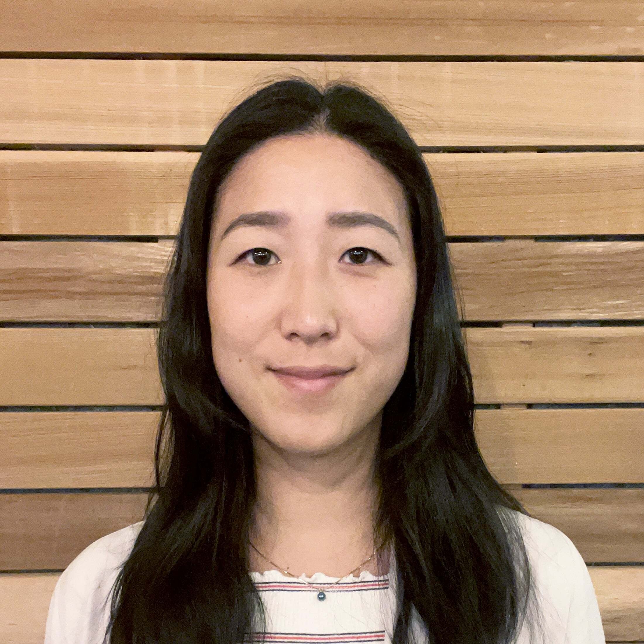 A woman with long black hair and a slight smile standing in front of a wooden wall.
