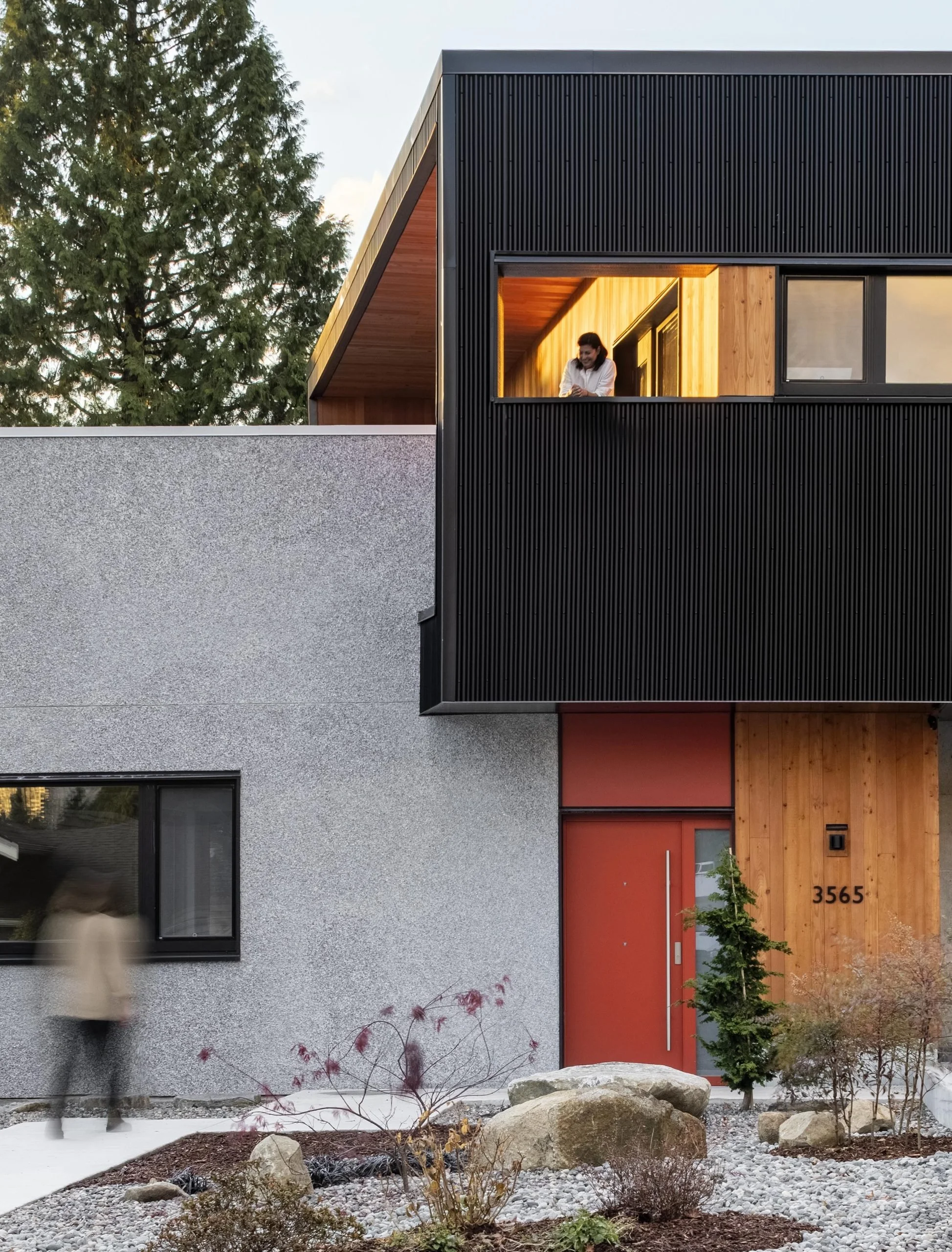 Modern house with a gray stucco lower level, red front door, wooden accents, and a black upper section. A woman stands at a second-floor window, and a blurred person is walking by in front.