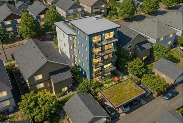 Aerial view of a modern apartment building with balconies, surrounded by houses and trees in a suburban neighborhood.