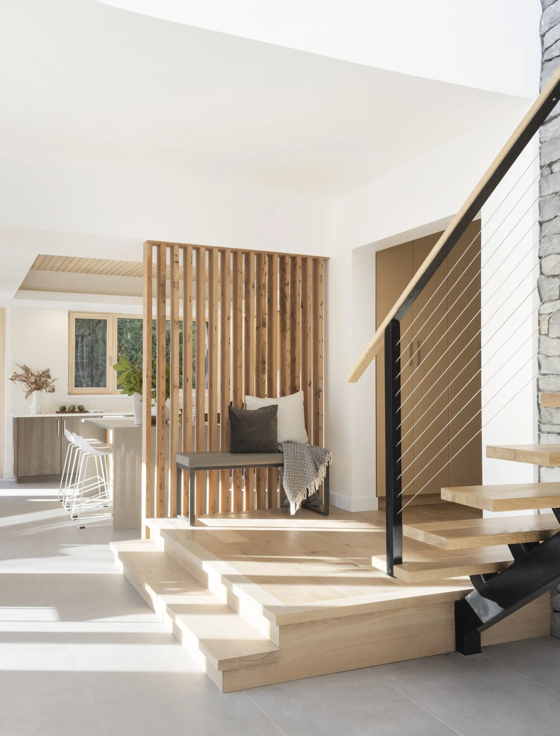Interior view of a modern home featuring a staircase with wooden steps and black railing, a wooden slat room divider, and a small sitting area with cushions and a blanket.