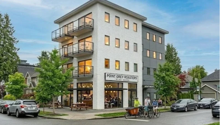 A four-story modern mixed-use building with retail space on the ground floor and apartments above, located on a city street with trees, parked cars, and pedestrians.