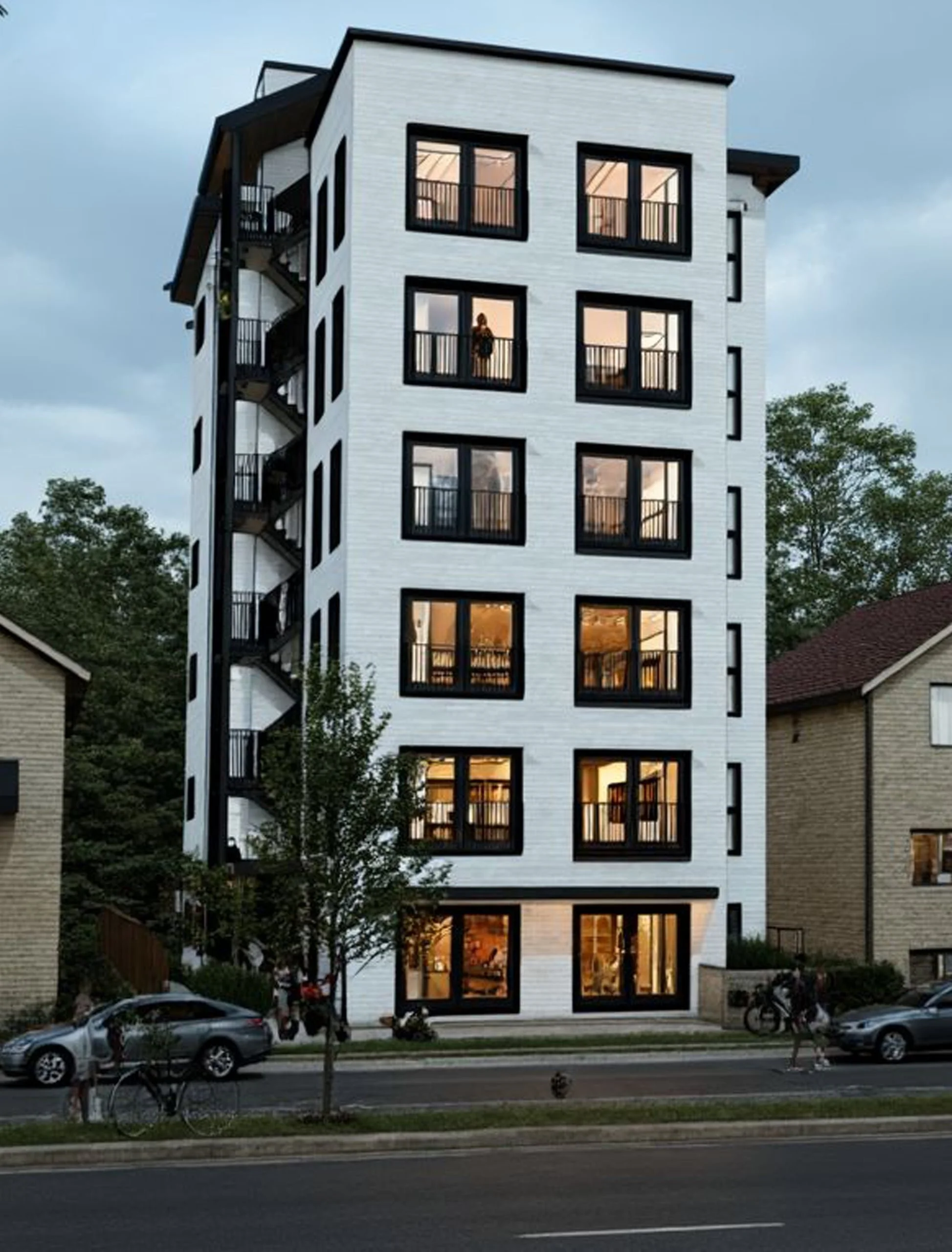 Multi-story modern white apartment building with large black frames on the windows, surrounded by trees and neighboring houses, with people walking and cars parked nearby.