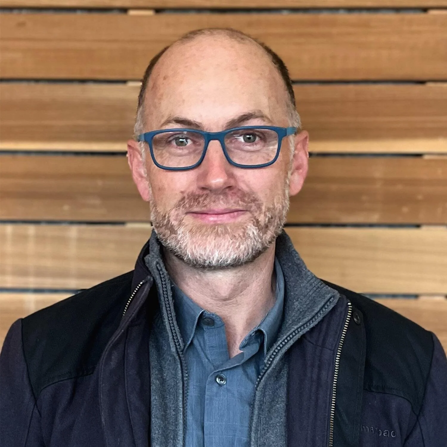 A middle-aged man with glasses, a beard, and a bald head, wearing a jacket and a collared shirt, standing in front of a wooden wall.