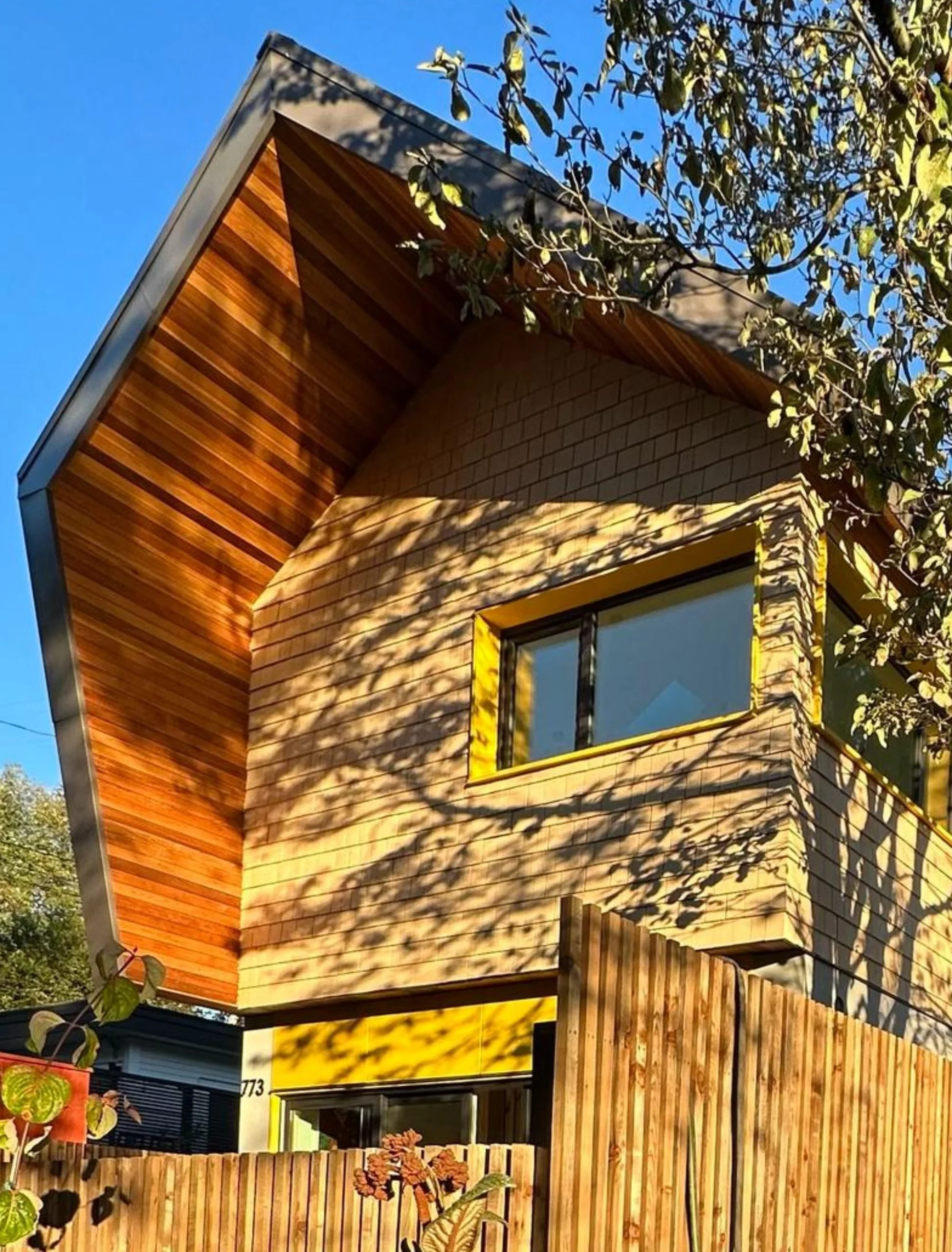 A modern house with wooden siding and a sloped roof, surrounded by trees and a wooden fence, under a blue sky.