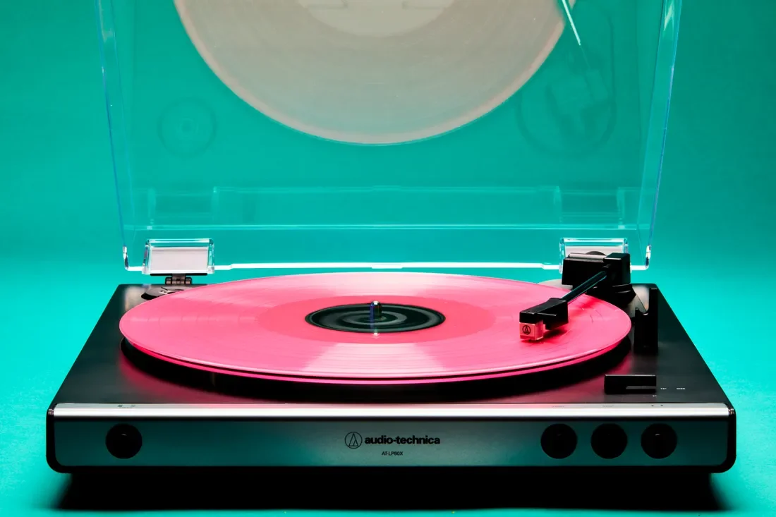 A pink vinyl record spinning on a black and silver Audio-Technica turntable with a clear plastic dust cover, against a teal background.