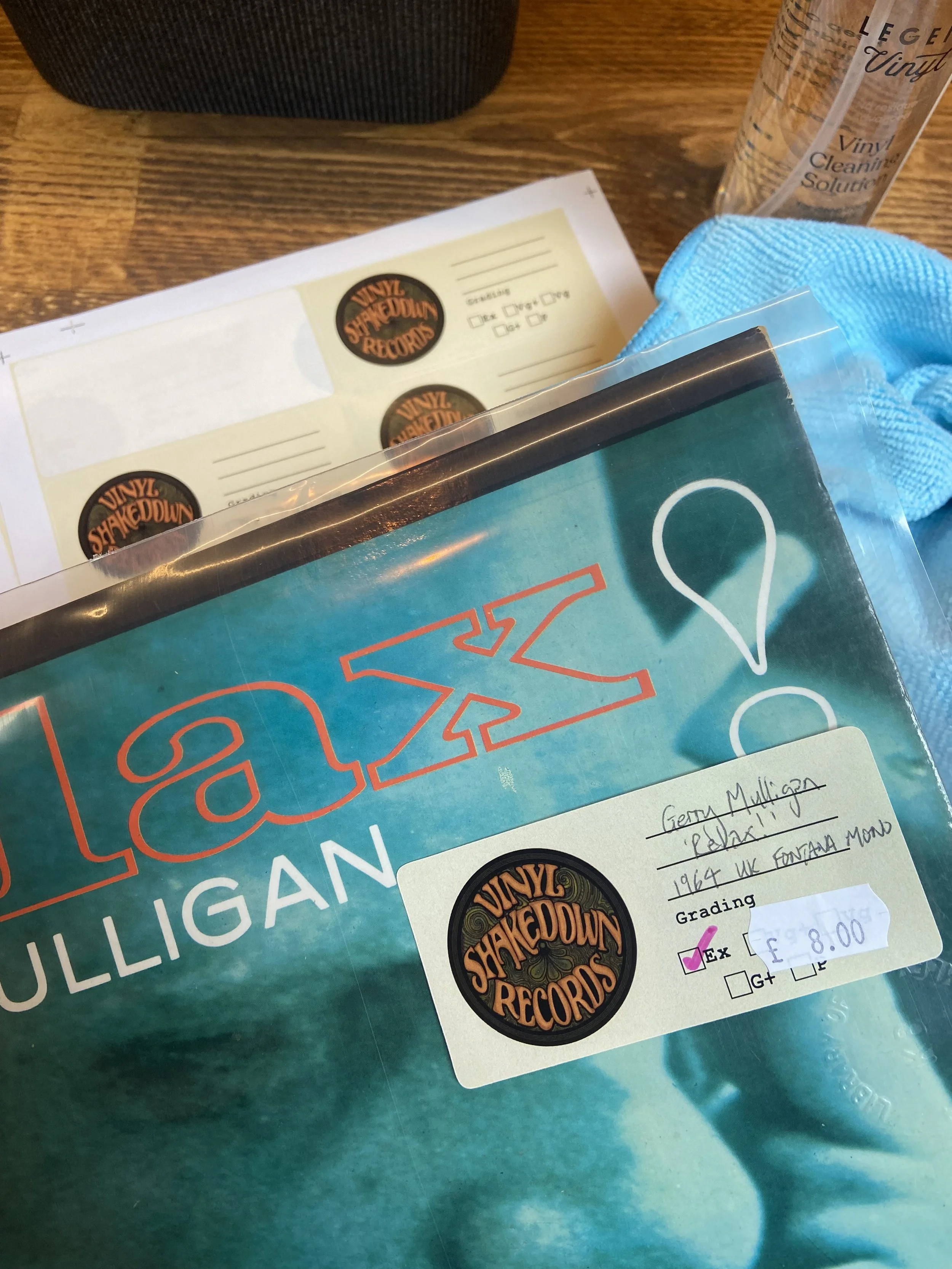 Close-up of vinyl records, a blue cloth, and a sticker with the Vinyl Shakedown Records logo on a wooden table.