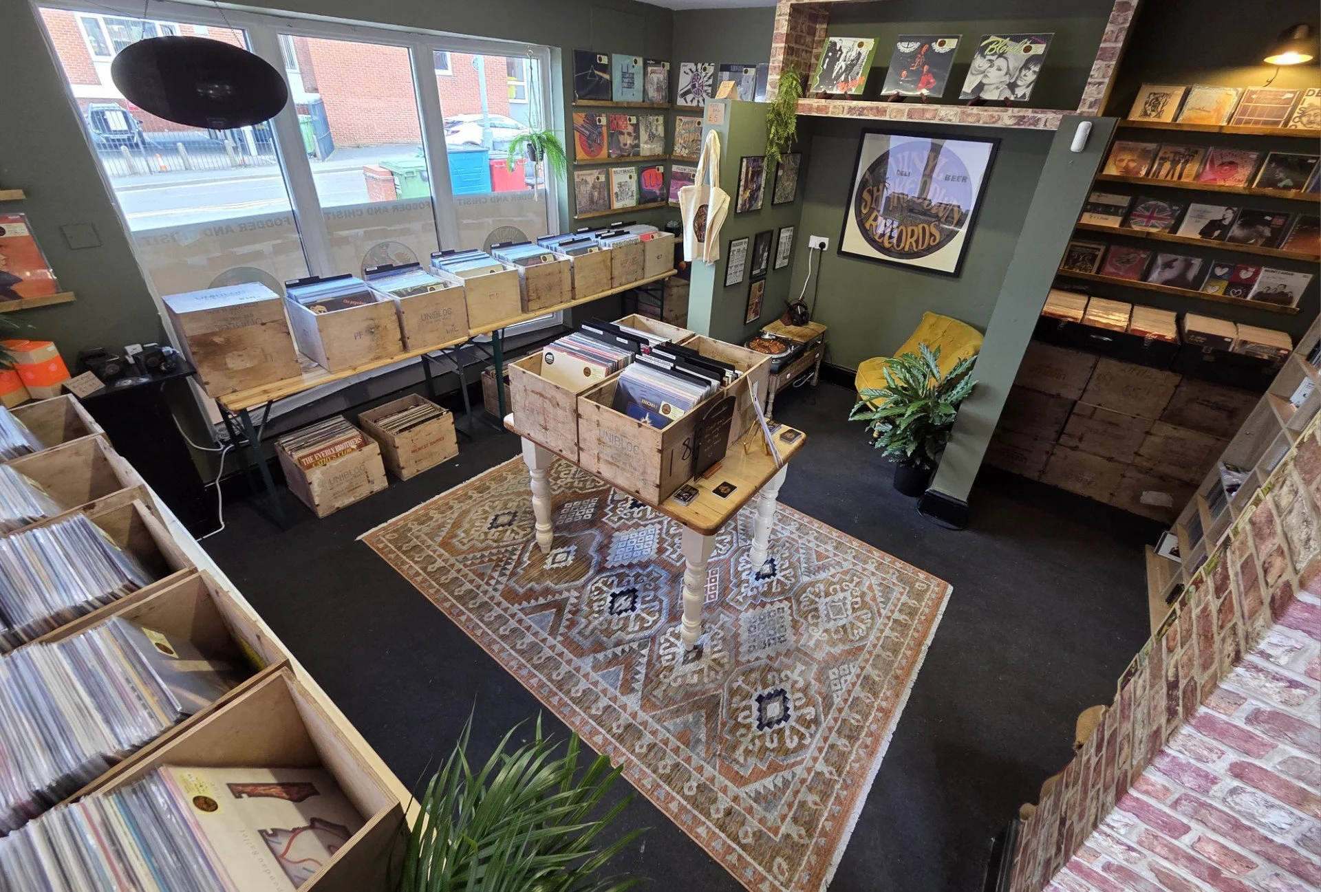 Inside a record store with wooden crates filled with vinyl records, a patterned rug on the floor, green walls decorated with album covers, a large window, and a brick accent wall.