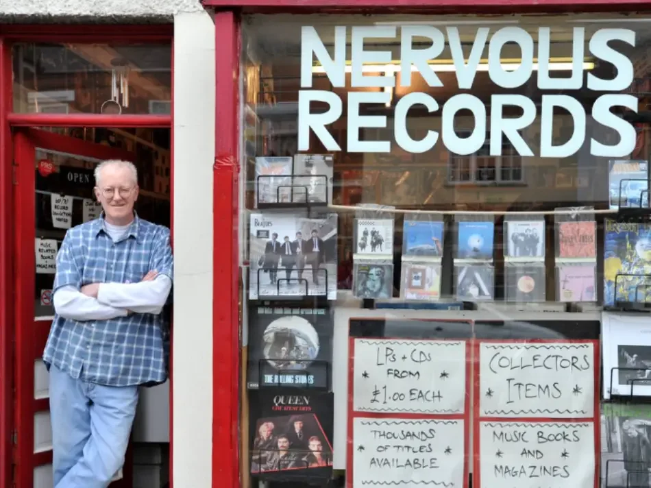 A man standing with arms crossed in front of a record store named Nervous Records filled with vinyl records.