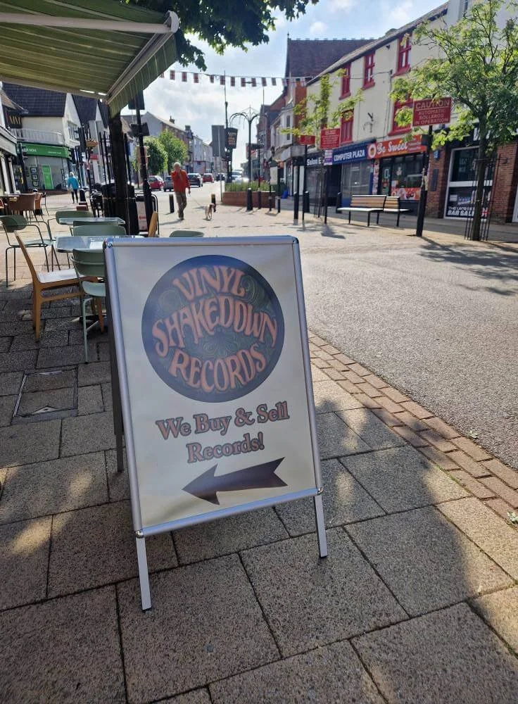 Street scene with a sidewalk, outdoor seating, and a sign for Vinyl Shakedown Records that says, "We Buy & Sell Records!" with an arrow pointing left.