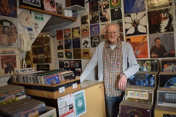 An elderly man with glasses, wearing a plaid shirt and a light-colored jacket, standing inside a record store surrounded by vinyl records on the shelves and wall.