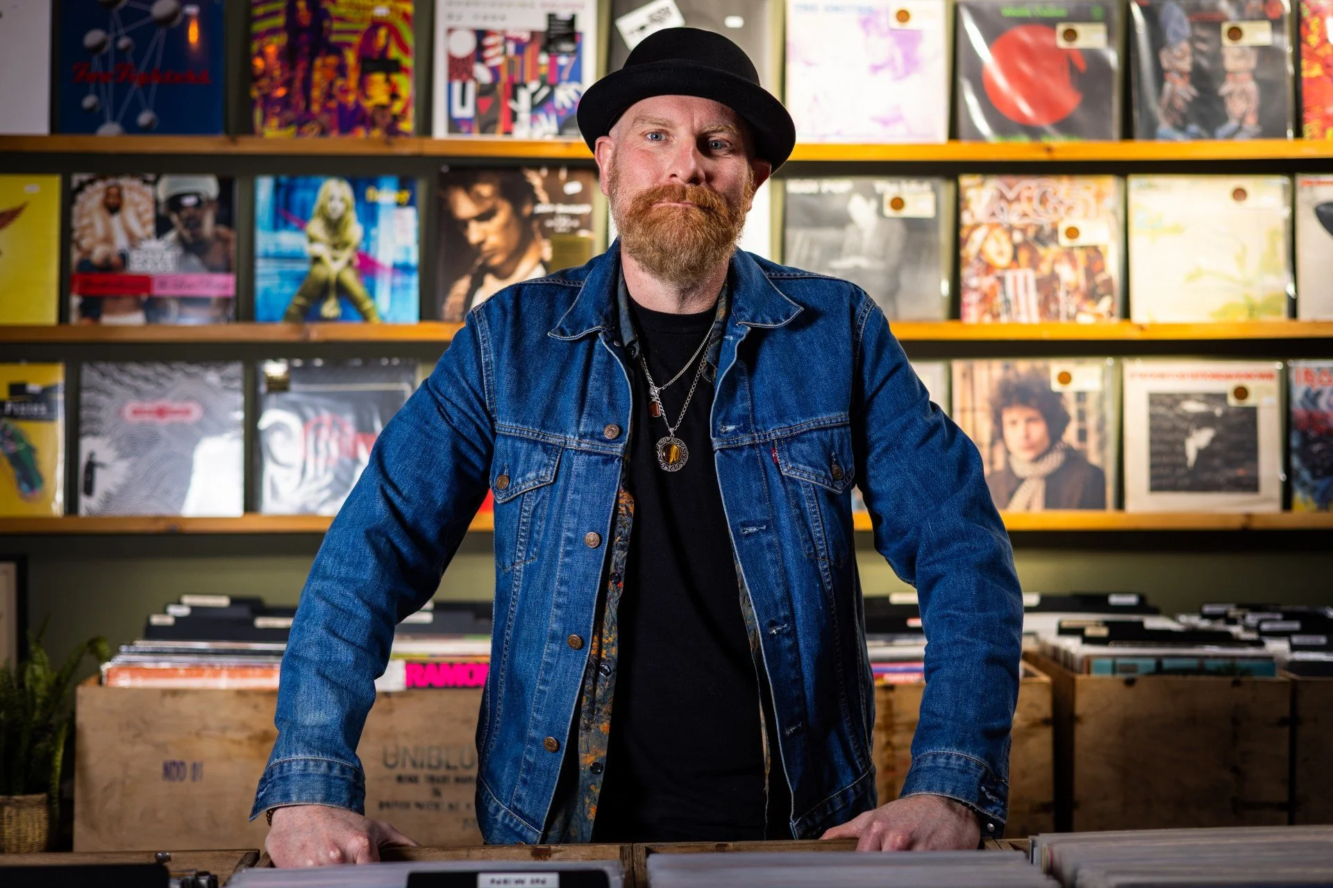 A man with a beard and mustache wearing a black hat, denim jacket, and layered necklaces, stands behind a record store counter with vinyl records display in the background.