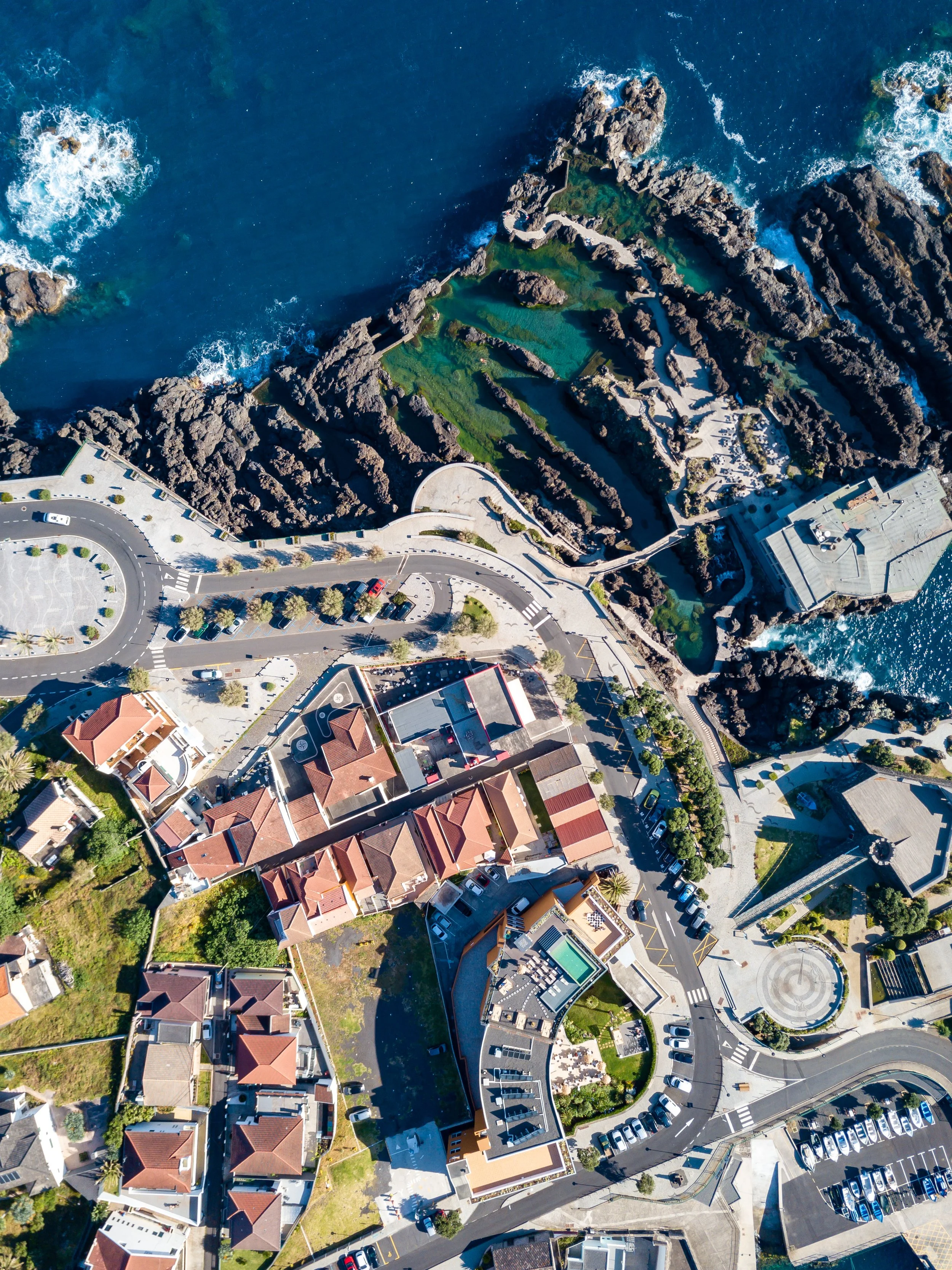 Aerial view of rocky coastline with green tide pools, surrounded by cars, roads, and residential buildings.