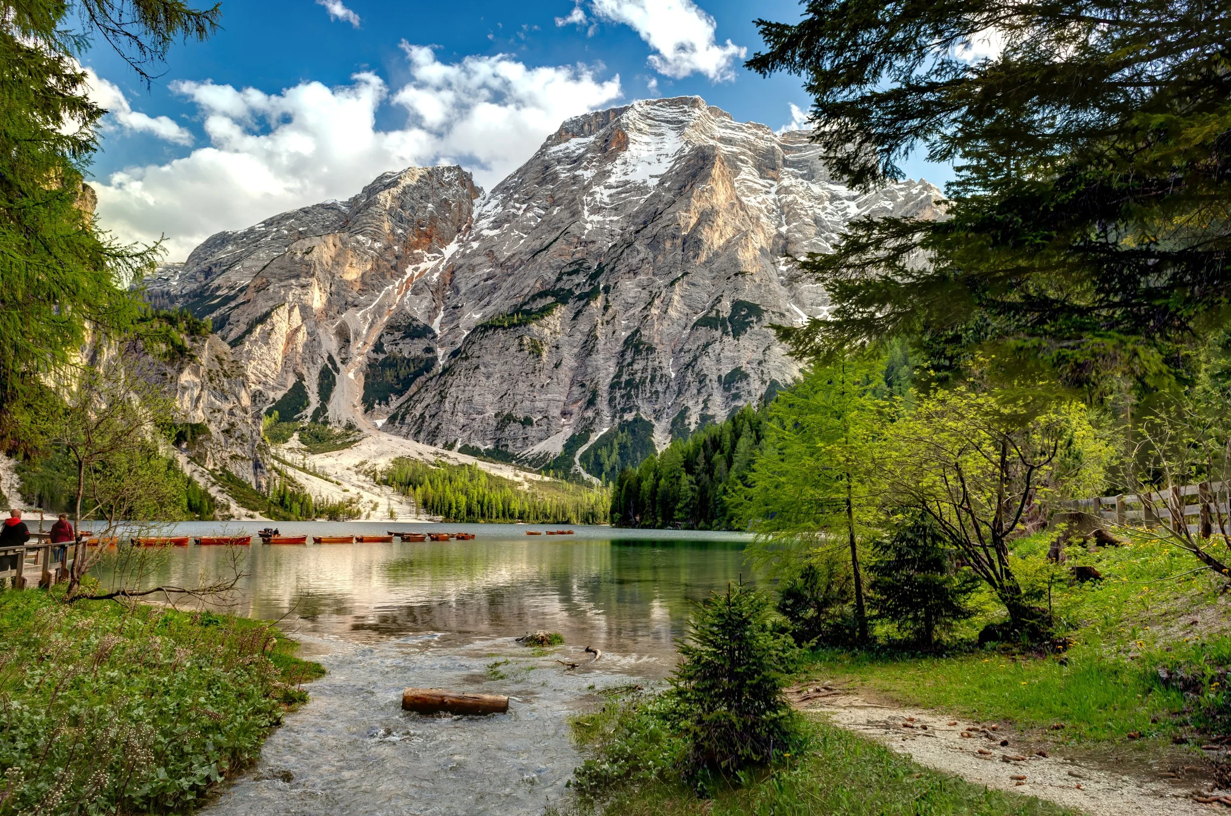 A scenic view of a mountain with snow at the peak, a calm lake, green trees, and a clear sky with some clouds.